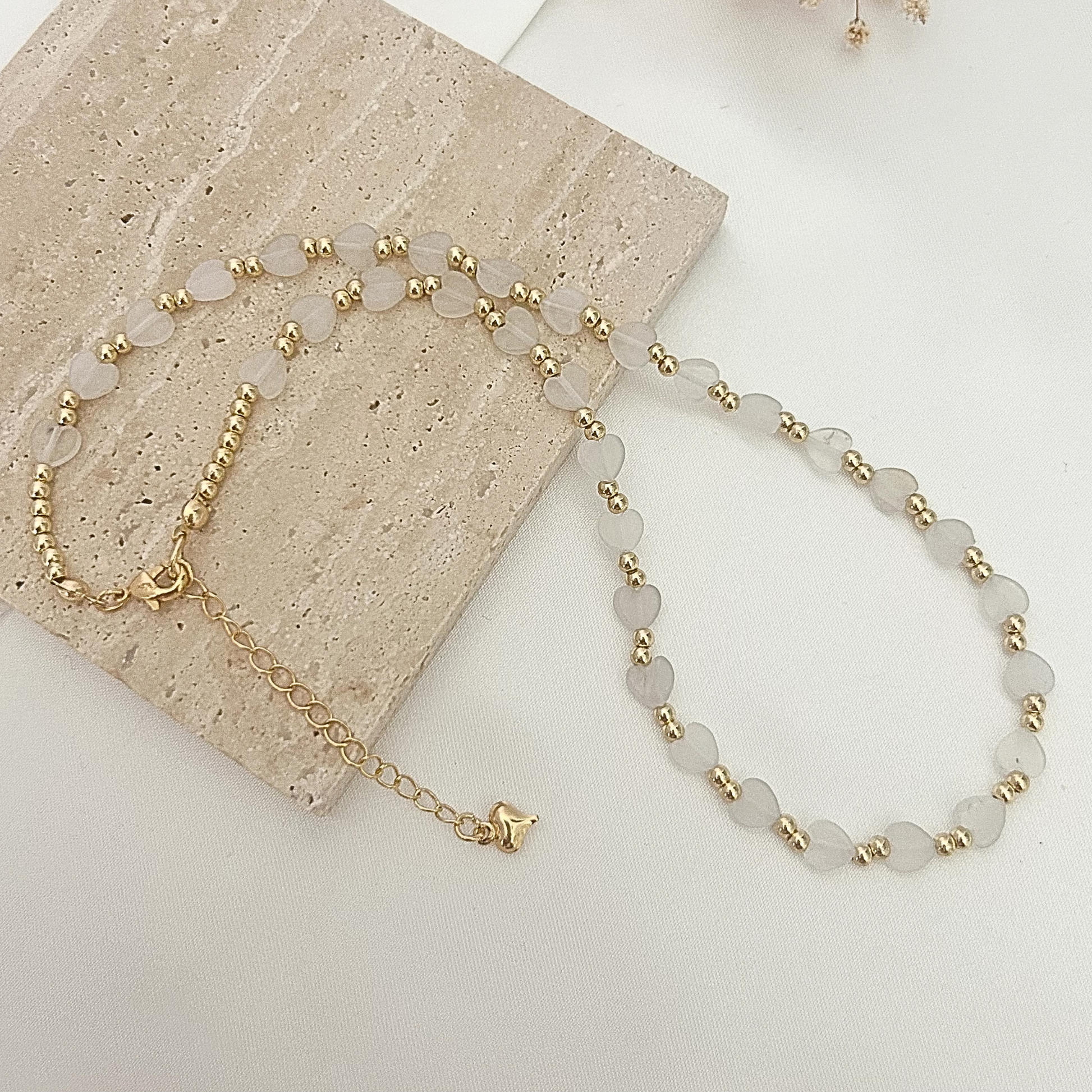 Delicate necklace featuring frosted heart-shaped beads paired with gold-tone spacer beads, displayed on a neutral stone surface.