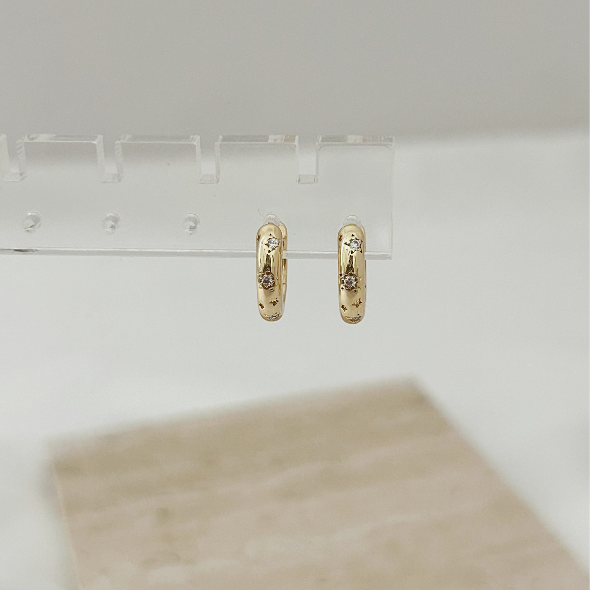 A pair of small gold hoop earrings with tiny clear stones, hanging from a clear display stand against a soft white background.