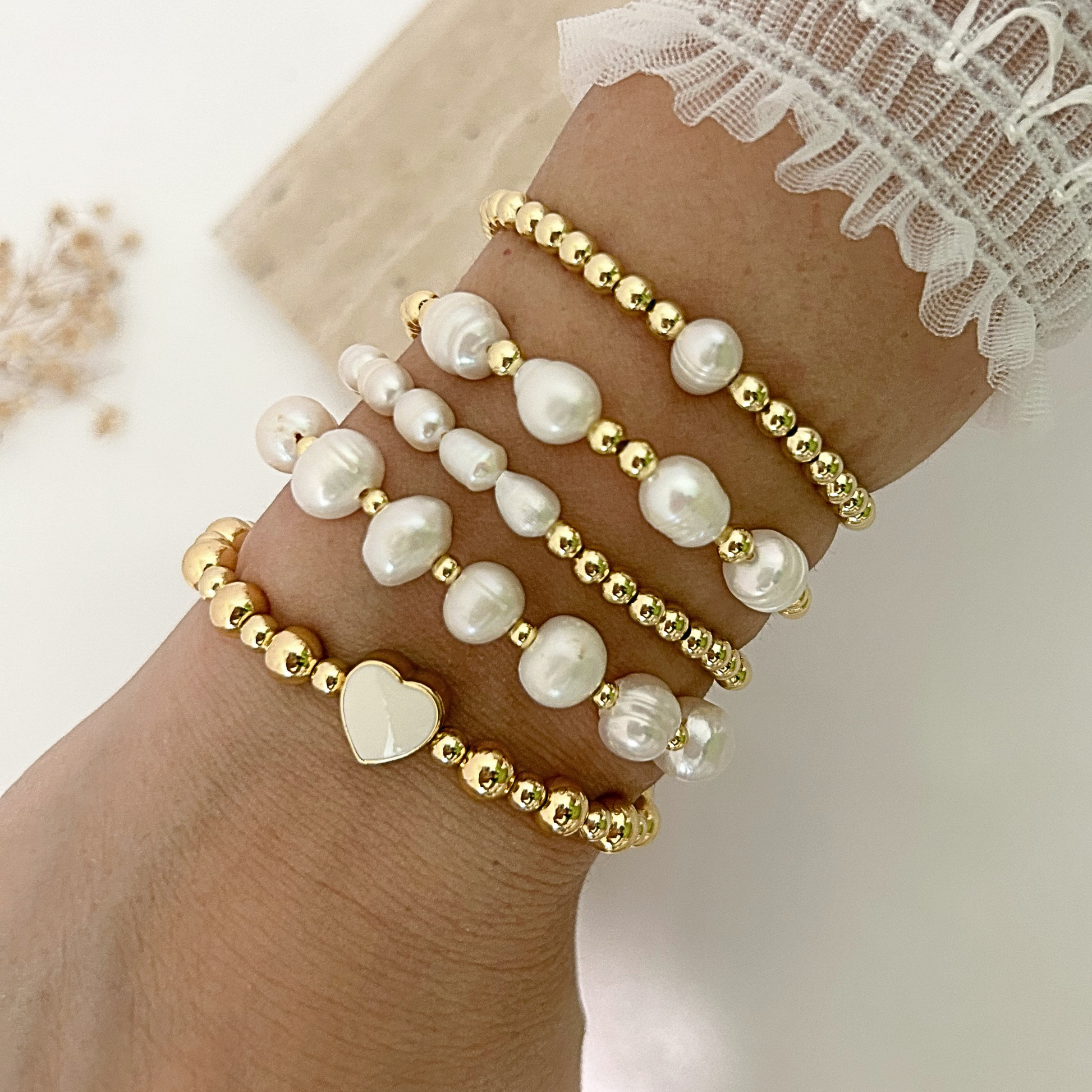 A close-up of a person's wrist wearing five layered beaded bracelets. The bracelets feature white freshwater pearls in irregular baroque shapes and polished gold-tone beads, with one bracelet displaying a white heart-shaped pearl.