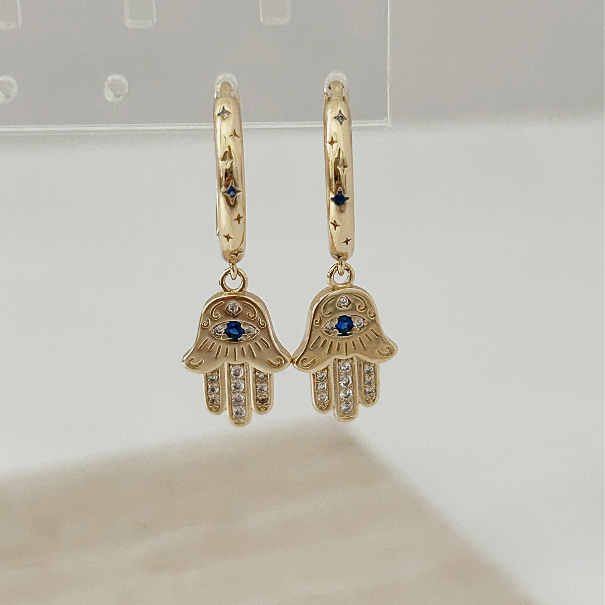 A pair of gold hamsa hand drop earrings with blue and clear crystal details, displayed on a neutral light gray background.