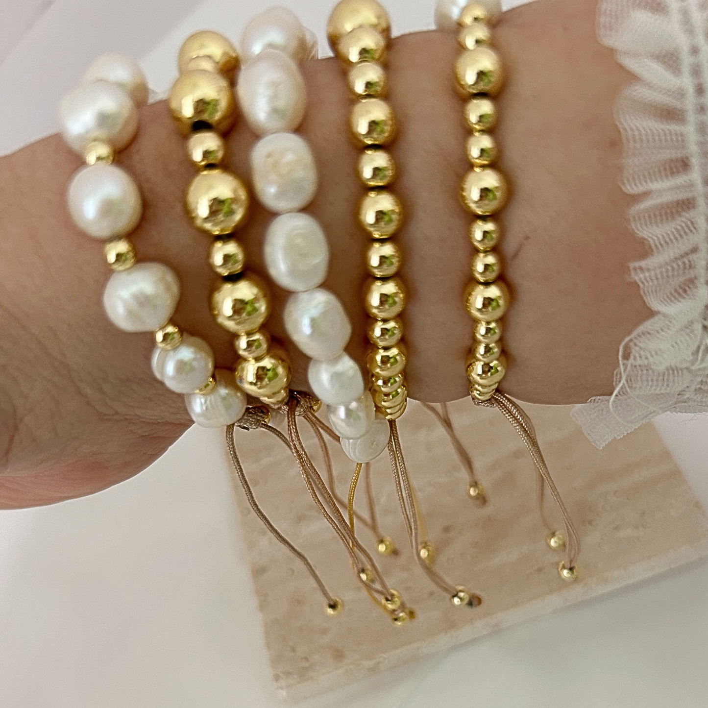A close-up of a wrist wearing five beaded bracelets: three strands with white irregular baroque pearls and two strands with shiny gold round beads, resting on a light wood block.