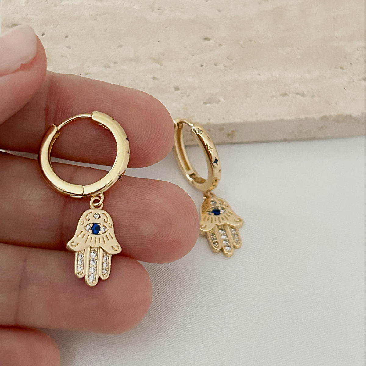 Gold huggie hoop earrings with dangling Hamsa hand charms, one held in a hand and the other resting beside it. The charms feature a blue gemstone for the eye and clear crystals along the fingers.