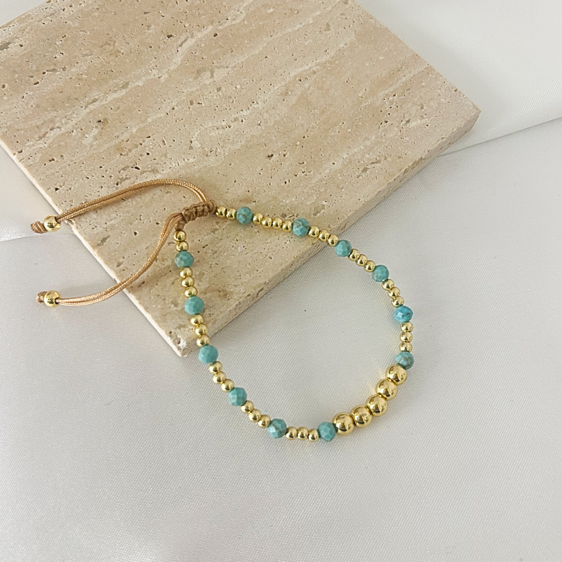 A beaded stretch bracelet with turquoise-blue beads and metallic gold spacer beads, featuring an adjustable tan cord closure. The bracelet is styled on a light beige stone tile over a white surface.
