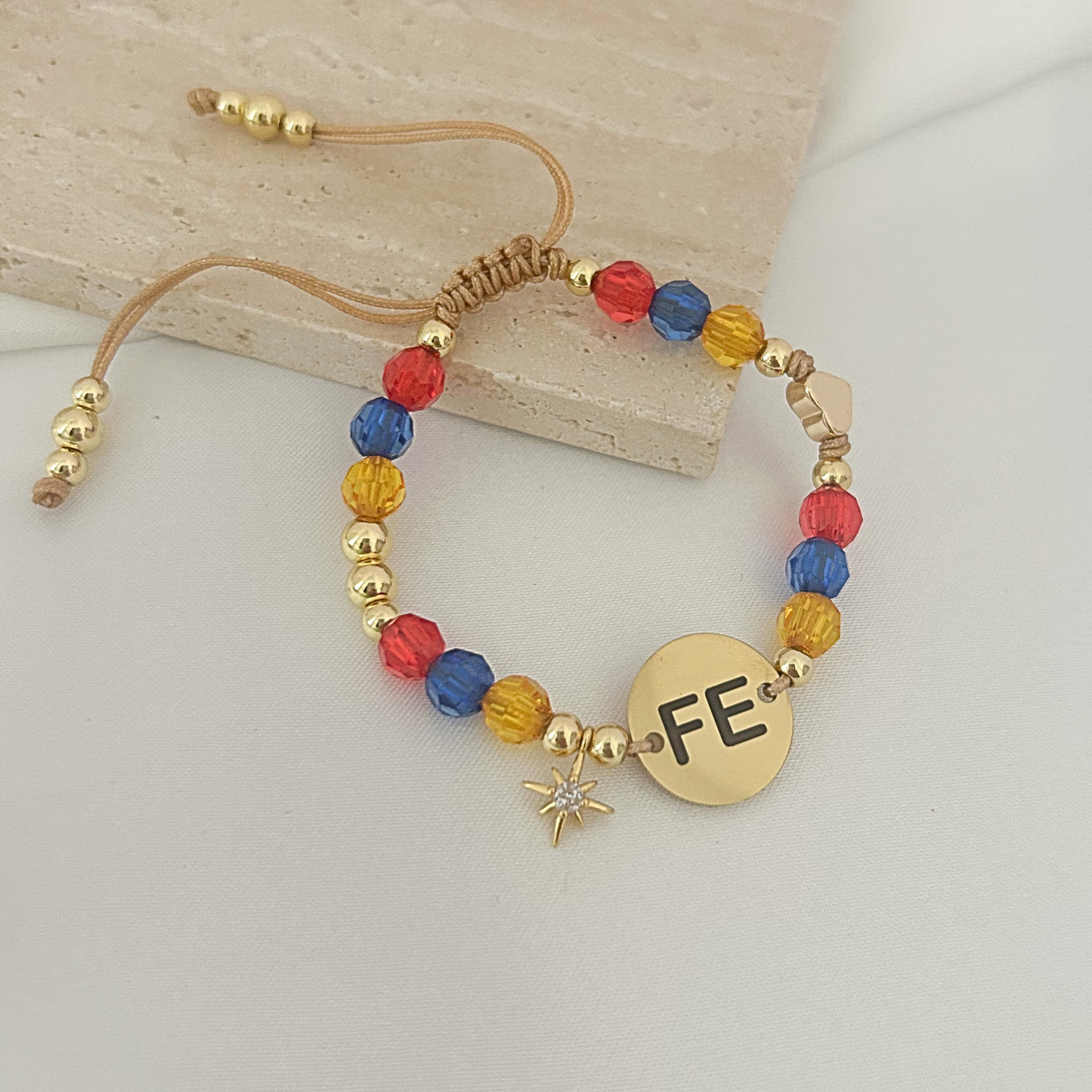 A gold-plated beaded bracelet with colorful red, blue, and yellow faceted beads, a round gold disc engraved with 'FE', and a small star charm, displayed on a light marble block over a white surface.