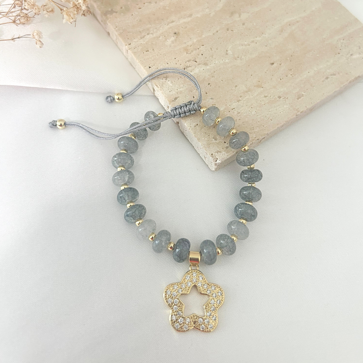 A beaded stretch bracelet featuring round gray stone beads, small gold spacer beads, a gray cord slider closure, and a gold-toned star-shaped pendant paved with clear rhinestones, displayed on a light surface with a stone prop.