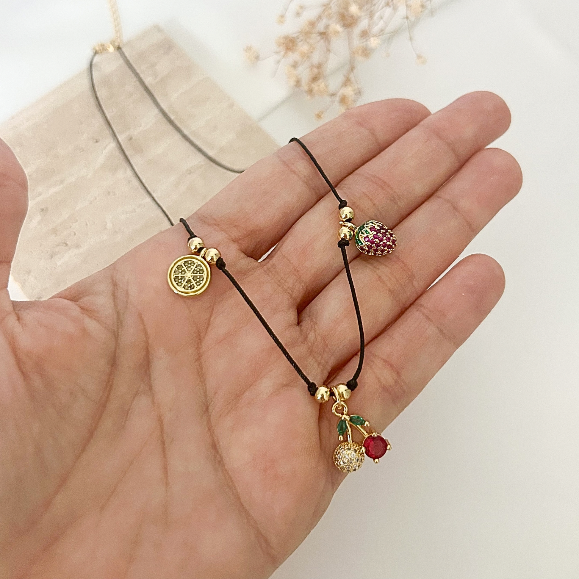 Close-up of a handmade charm necklace showcasing cherry, strawberry, watermelon, and lemon-inspired pendants on a minimalist black cord.