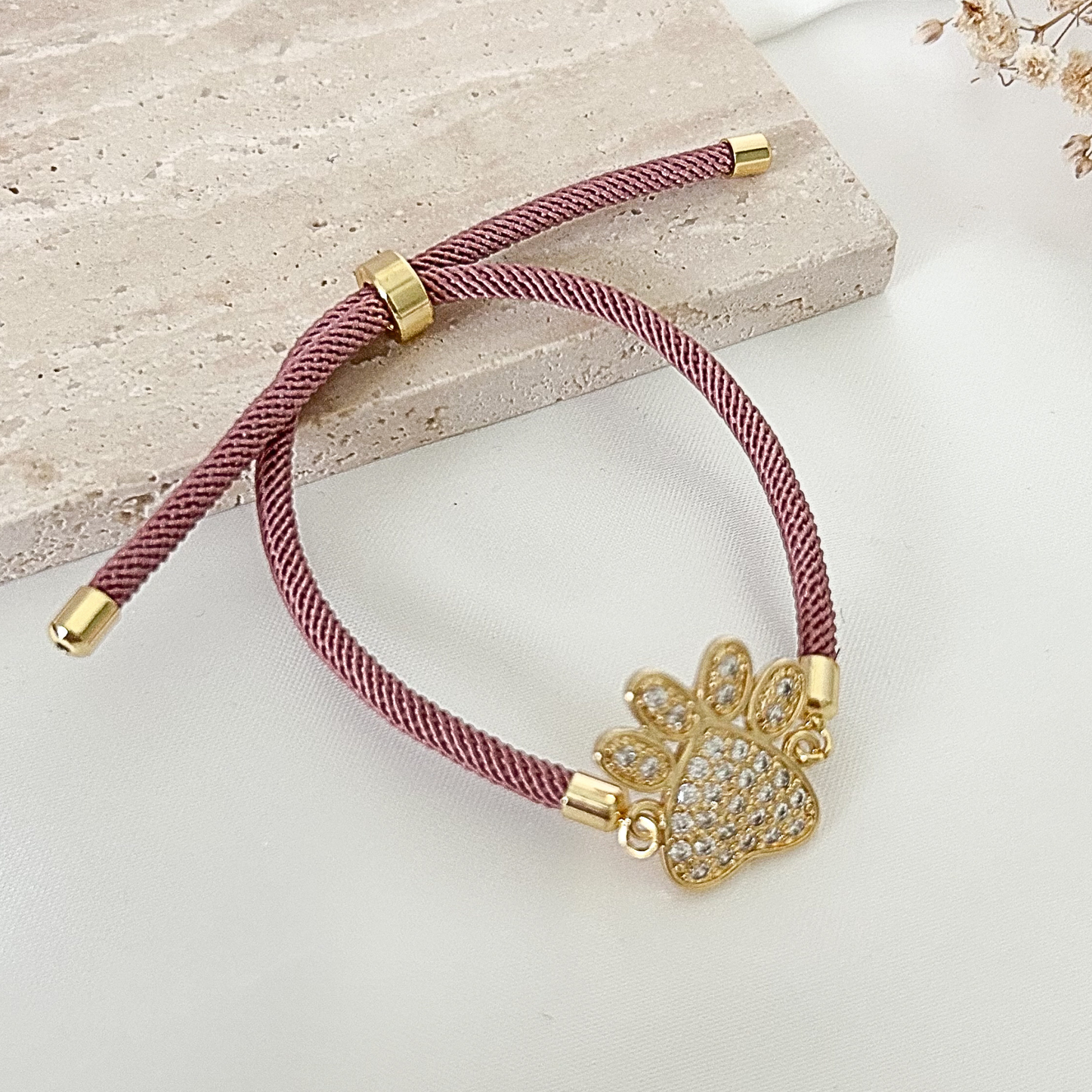 Adjustable pet lover bracelet featuring a sparkling paw charm with crystal accents, set on a dusty rose braided cord with gold-tone details against a soft neutral background.