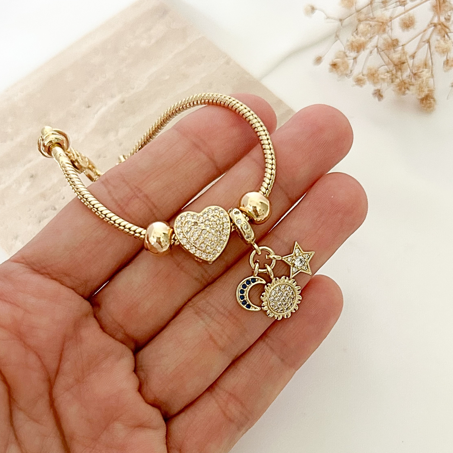 Close-up of a gold celestial charm bracelet held in hand, featuring a crystal-studded heart, sparkling sun charm, crescent moon with blue accents, and a delicate star detail.
