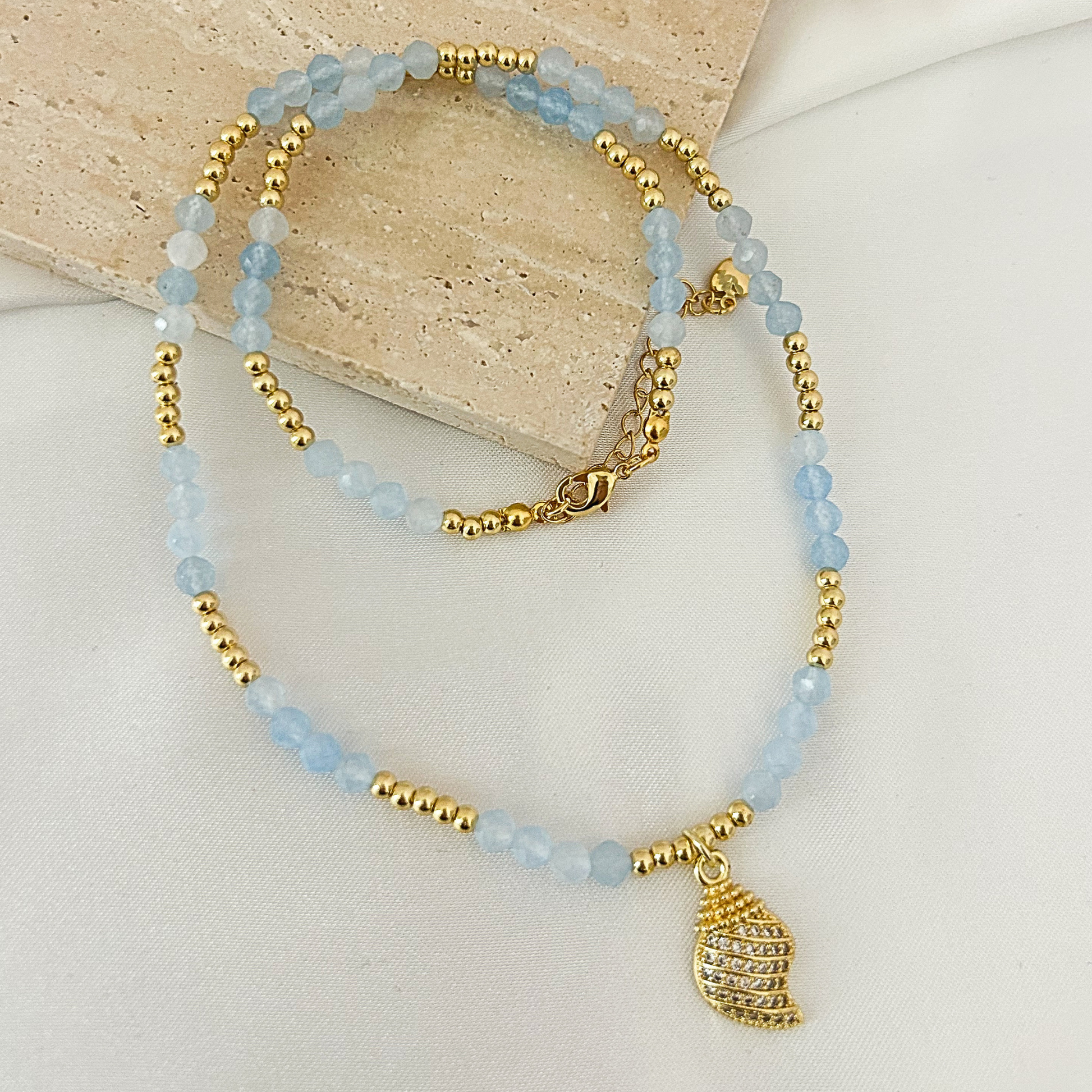 A beaded necklace with light blue faceted beads, gold spacer beads, and a gold seashell pendant, displayed on a neutral fabric background with a stone prop.