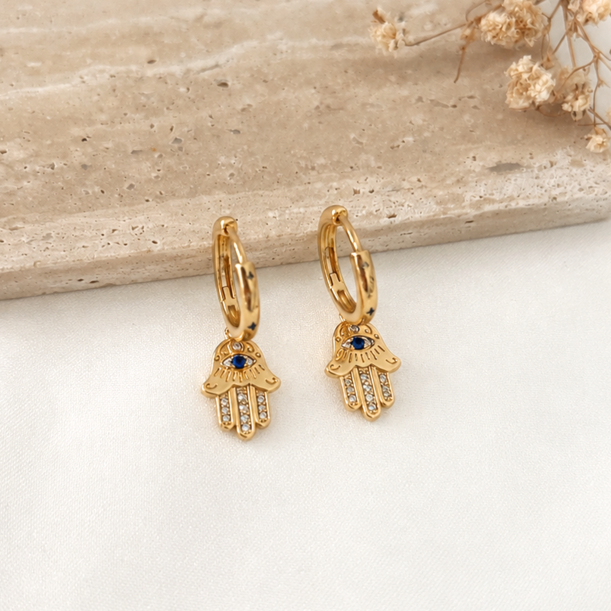 Pair of gold-tone huggie hoop earrings featuring dangling Hamsa hand charms with blue evil eye accents and sparkling cubic zirconia details. Meaningful protective earrings with a modern minimalist design, perfect for everyday wear or layering with other gold jewelry.