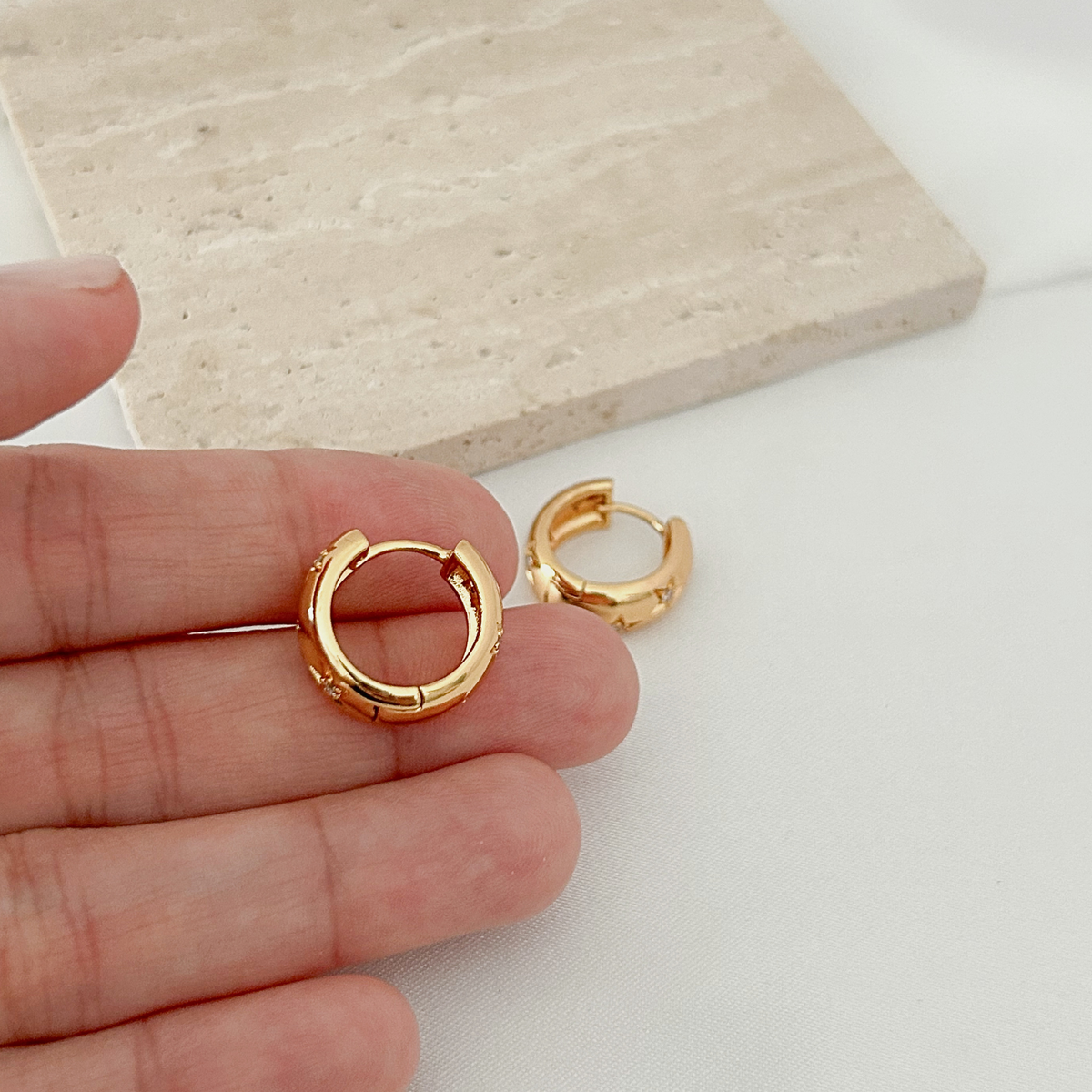 A pair of small gold huggie hoop earrings held between fingers, with one earring also resting on a white surface. A beige stone tile is blurred in the background.