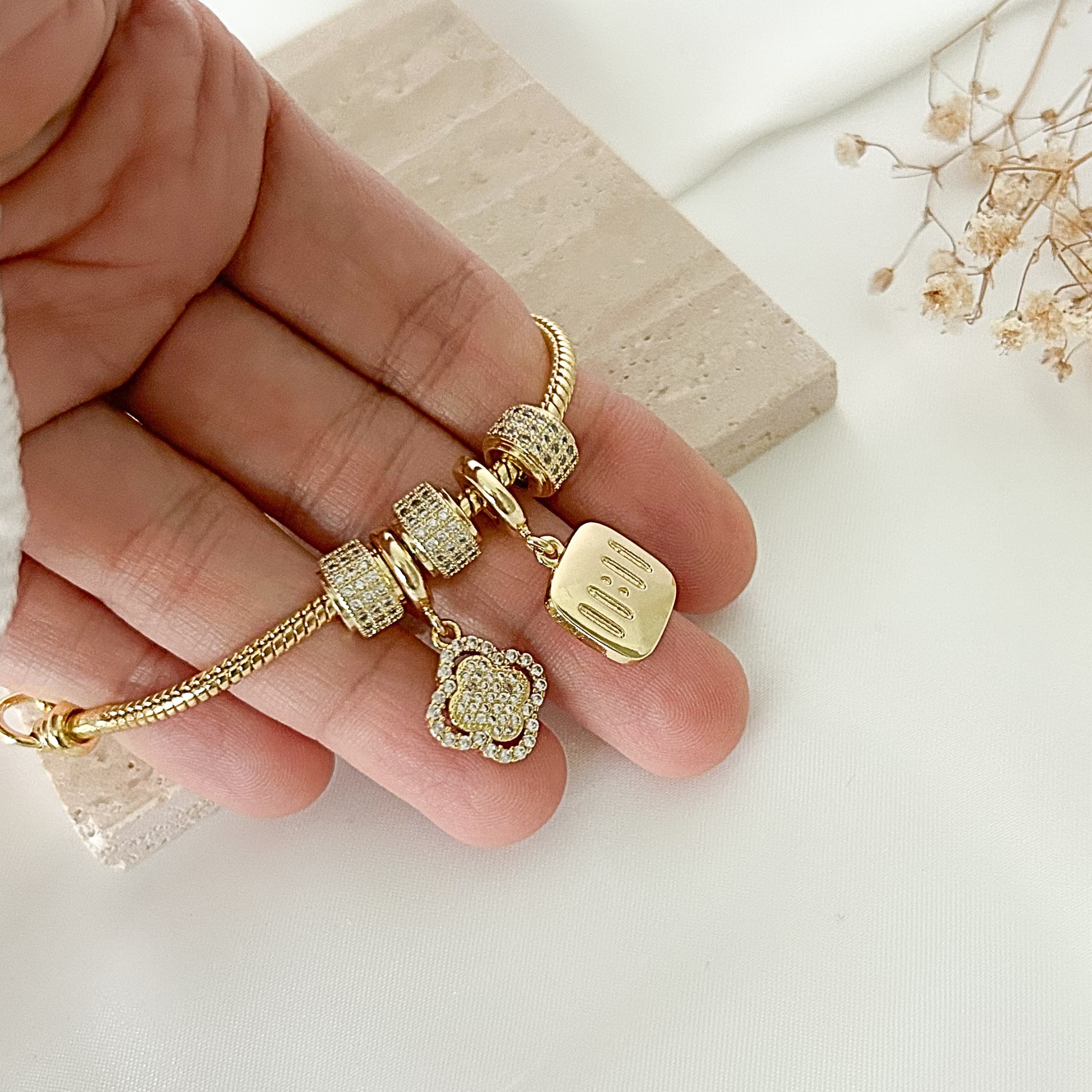 Close-up of a gold charm bracelet held in hand, showcasing shimmering pavé details, a floral-inspired charm, and a polished “mom” pendant, perfect for Mother’s Day or everyday wear.