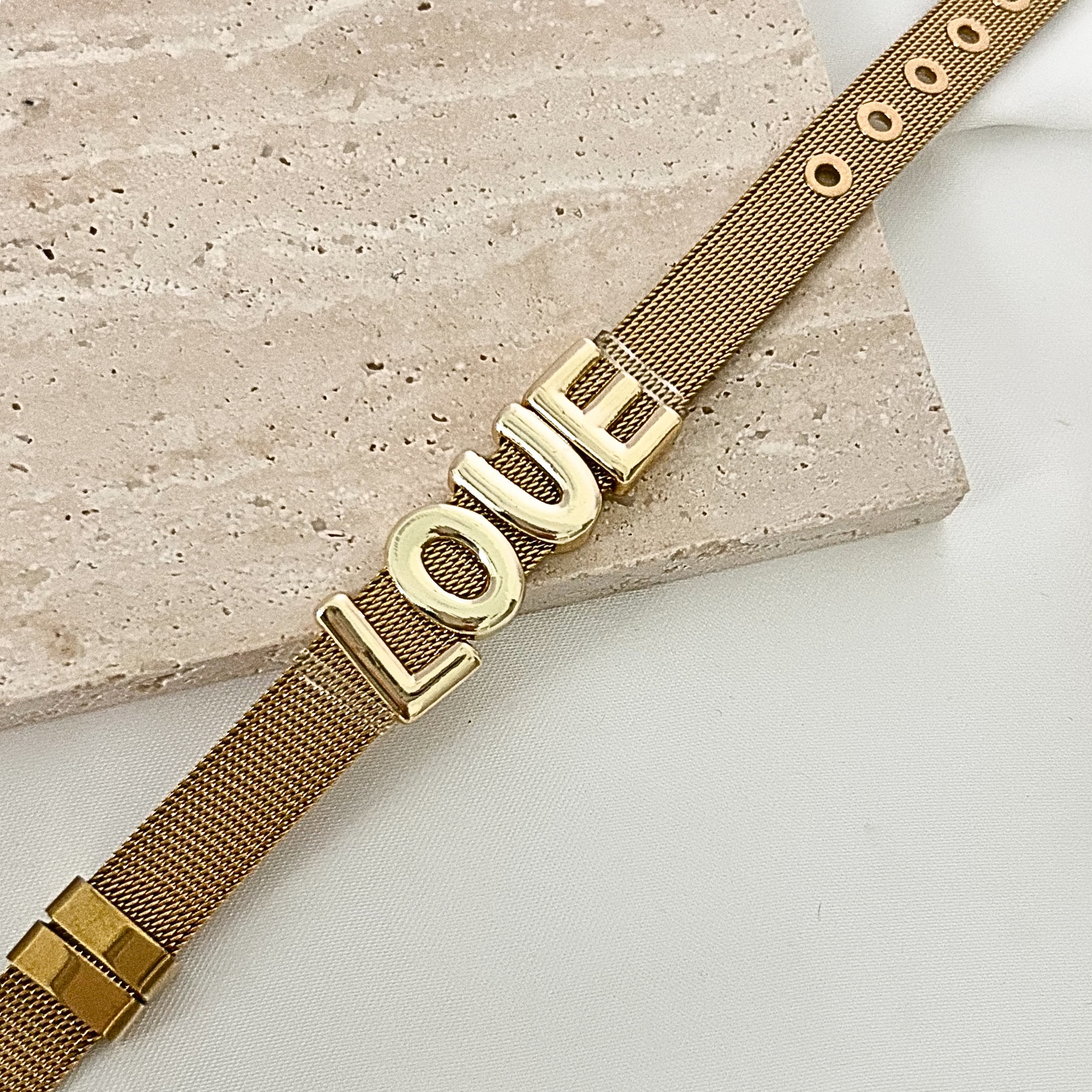 Elegant gold-tone stainless steel mesh bracelet with oversized LOVE letters, styled on a light stone surface for a modern and minimalist look.