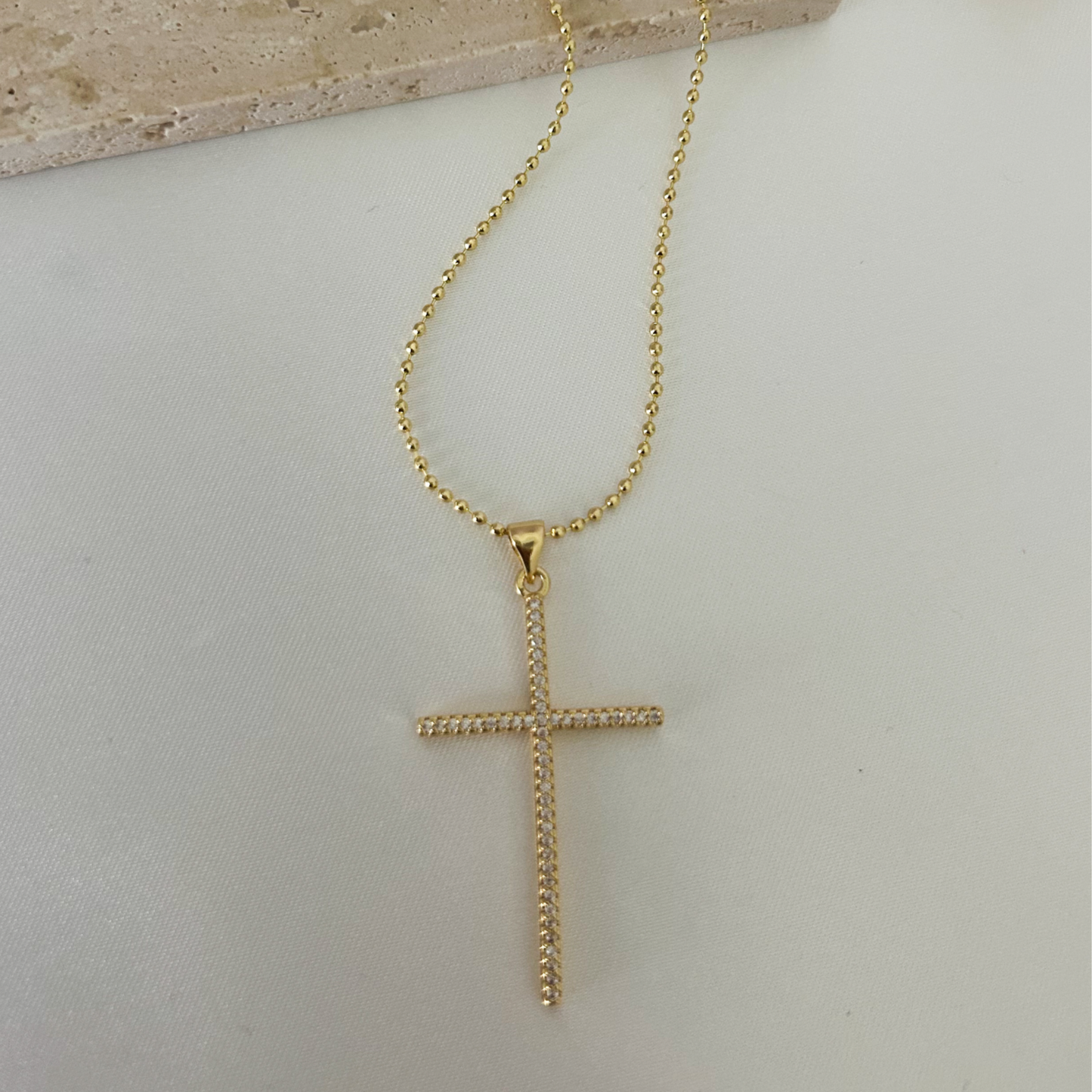 Primer plano de collar dorado con dije de cruz de cristales sobre fondo neutro y elegante, ideal para looks de fe y estilo.