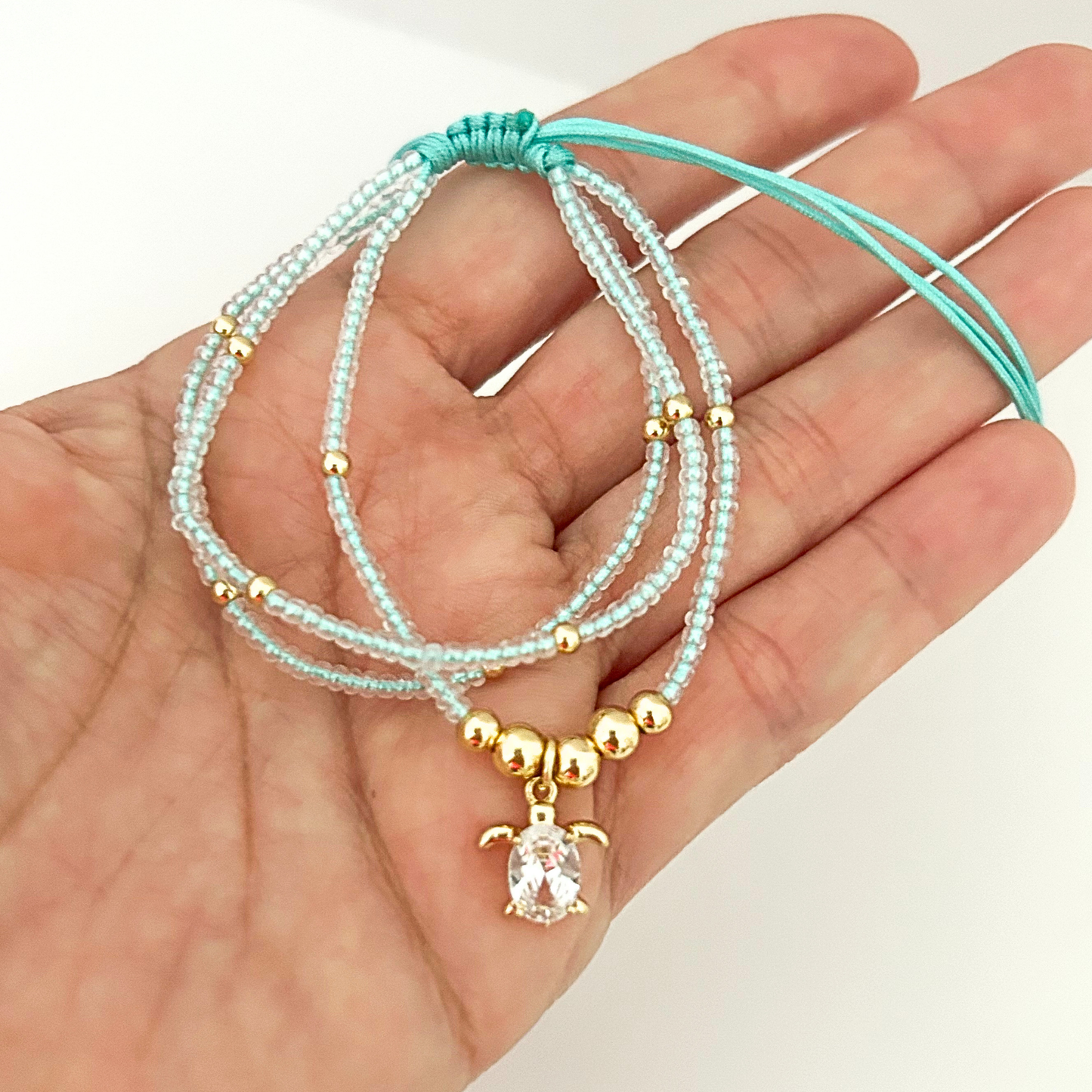 Handmade ocean-inspired bracelet featuring light aqua seed beads, gold-tone accents, and a crystal sea turtle centerpiece.