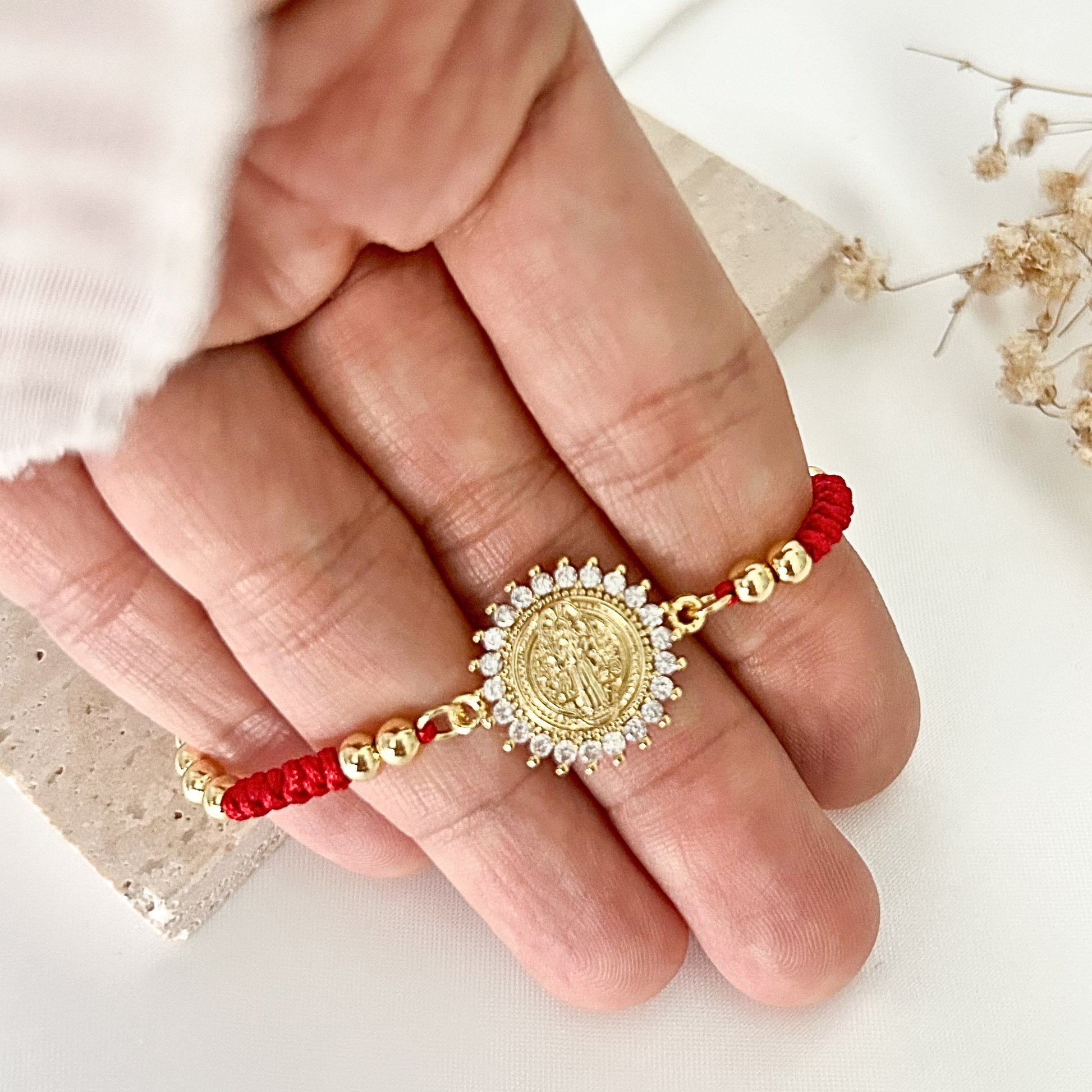 Close-up of a handcrafted Saint Benedict bracelet combining vibrant red cord, polished gold-tone beads, and a crystal halo pendant.