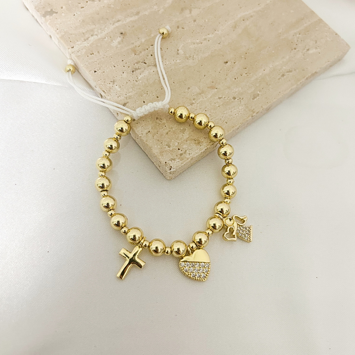 Gold beaded bracelet with cross, heart, and angel charms on a stone tray
