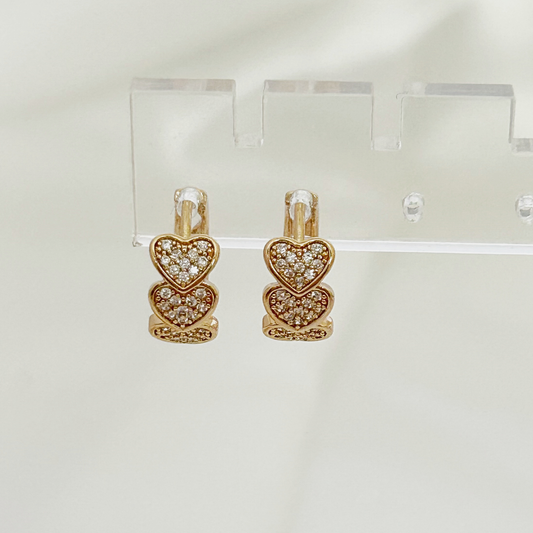 A pair of gold-toned heart-shaped drop earrings with pavé-set clear stones, displayed on a clear acrylic stand against a soft white background.