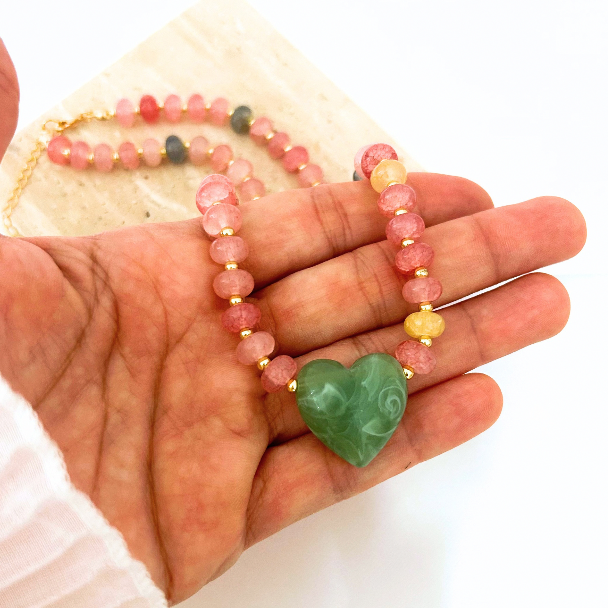 Close-up of a handcrafted necklace showcasing faceted pink, gray, and golden-yellow gemstone beads separated by gold spacers, finished with a large carved green heart pendant. A unique artisan-inspired jewelry piece with vibrant natural colors.