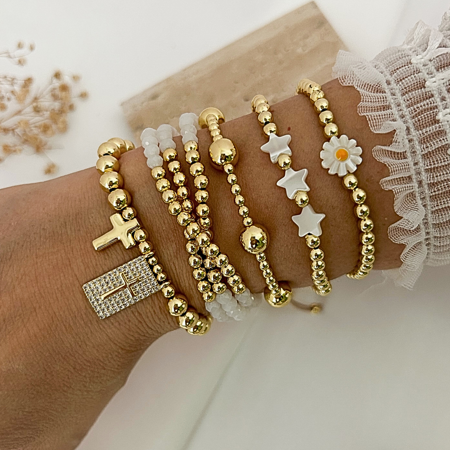 A close-up of a person's wrist wearing multiple delicate beaded bracelets. The bracelets include gold-tone beads, clear crystal beads, a gold cross charm, a gold rectangular crystal pendant, white star charms, a white daisy flower charm, and a gold ball charm.