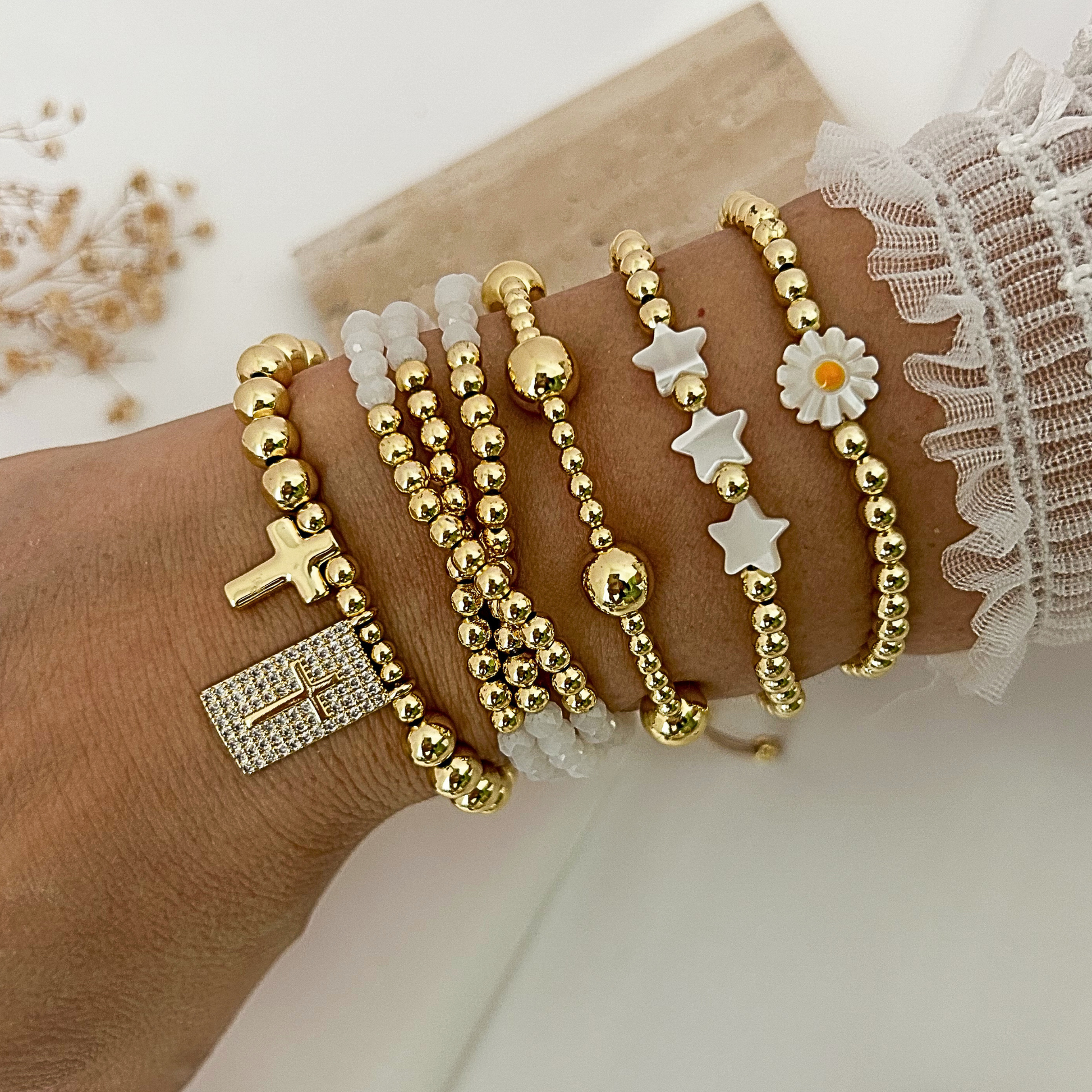 A close-up of a person's wrist wearing multiple delicate beaded bracelets. The bracelets include gold-tone beads, clear crystal beads, a gold cross charm, a gold rectangular crystal pendant, white star charms, a white daisy flower charm, and a gold ball charm.