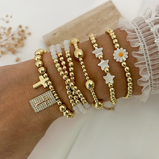 A close-up of a person's wrist wearing multiple delicate beaded bracelets. The bracelets include gold-tone beads, clear crystal beads, a gold cross charm, a gold rectangular crystal pendant, white star charms, a white daisy flower charm, and a gold ball charm.