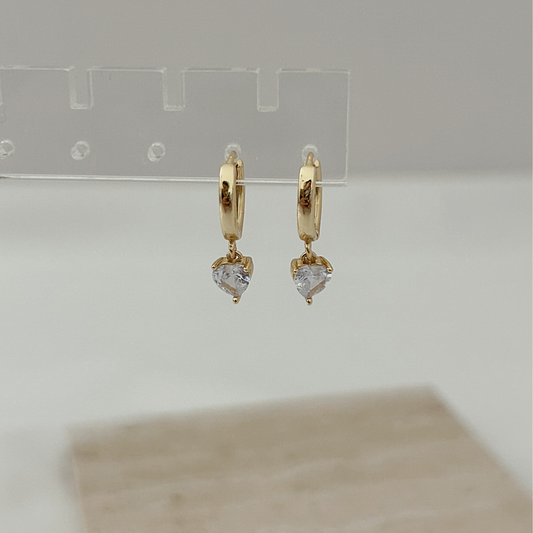 A pair of small gold huggie hoop earrings with dangling heart-shaped clear stones, displayed on a transparent stand against a soft light gray background.