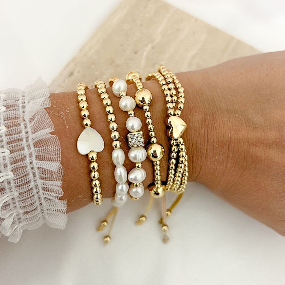 Close-up of layered gold beaded bracelets with freshwater pearls, heart charms, and an adjustable design.