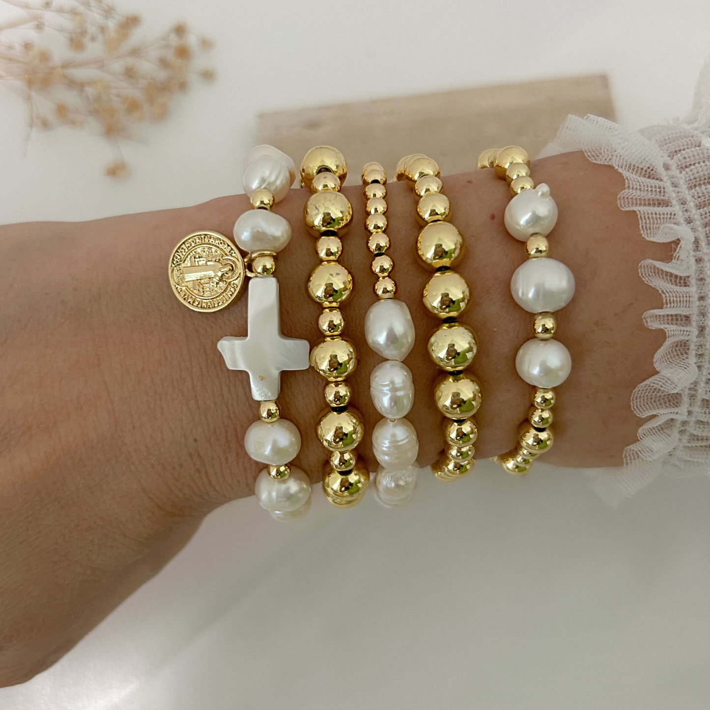 A close-up of a person's wrist wearing multiple beaded bracelets, including white pearl-like beads, gold-tone round beads, a white cross charm, and a round gold coin charm, with a white lace sleeve visible at the edge.