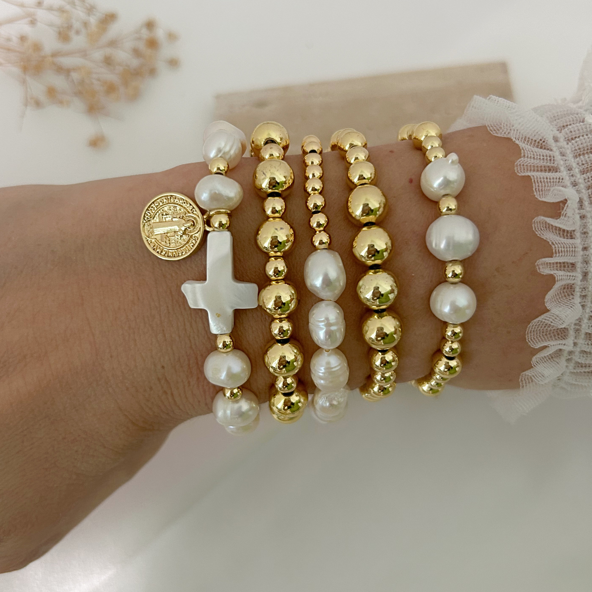 A close-up of a person's wrist wearing multiple beaded bracelets, including white pearl-like beads, gold-tone round beads, a white cross charm, and a round gold coin charm, with a white lace sleeve visible at the edge.