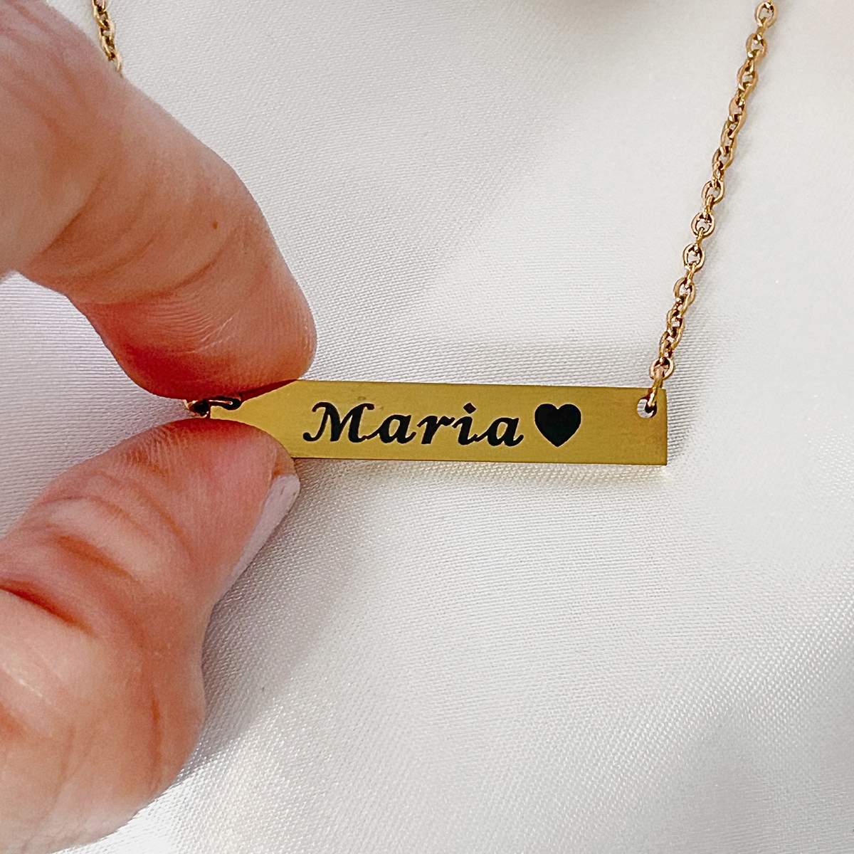 A close-up of a hand holding a gold bar necklace with the name 'Maria' and a black heart engraved on it, against a white fabric background.