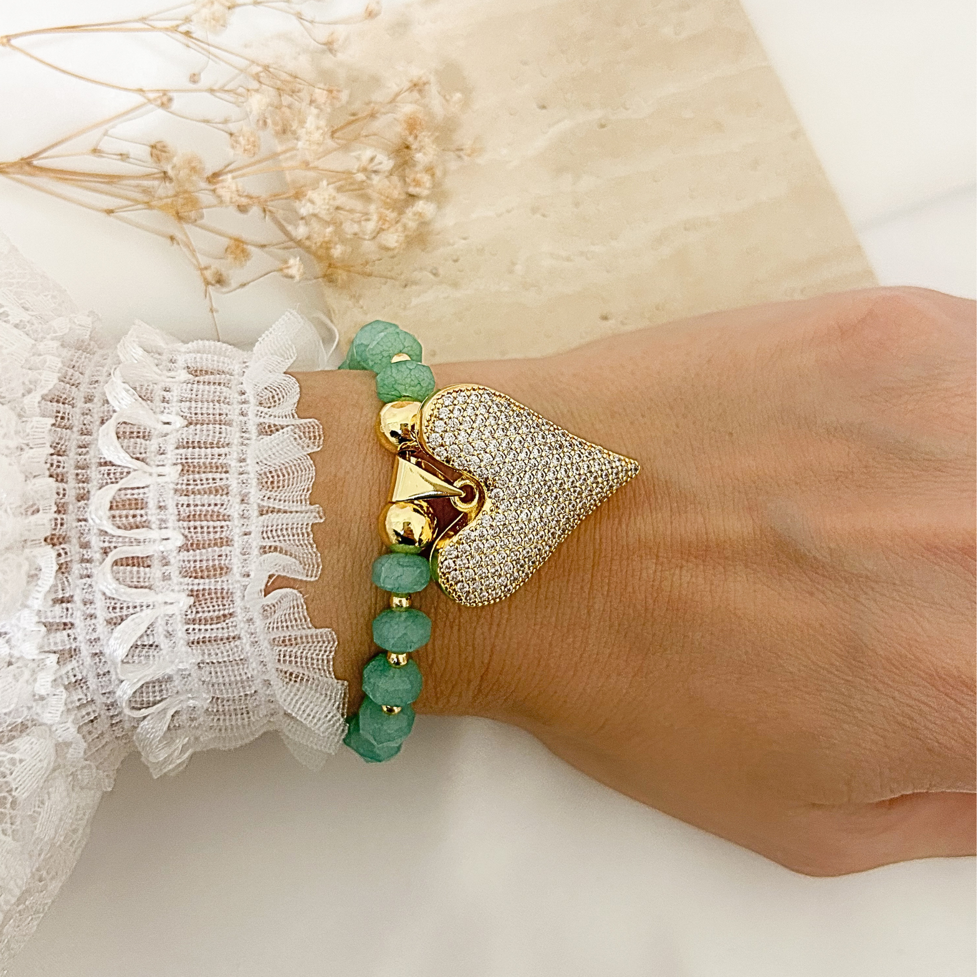 A close-up of a person's wrist wearing two bracelets: one with green faceted beads and one with a large gold-tone heart charm and a pavé-set silver heart, styled with a white lace sleeve against a light neutral background.