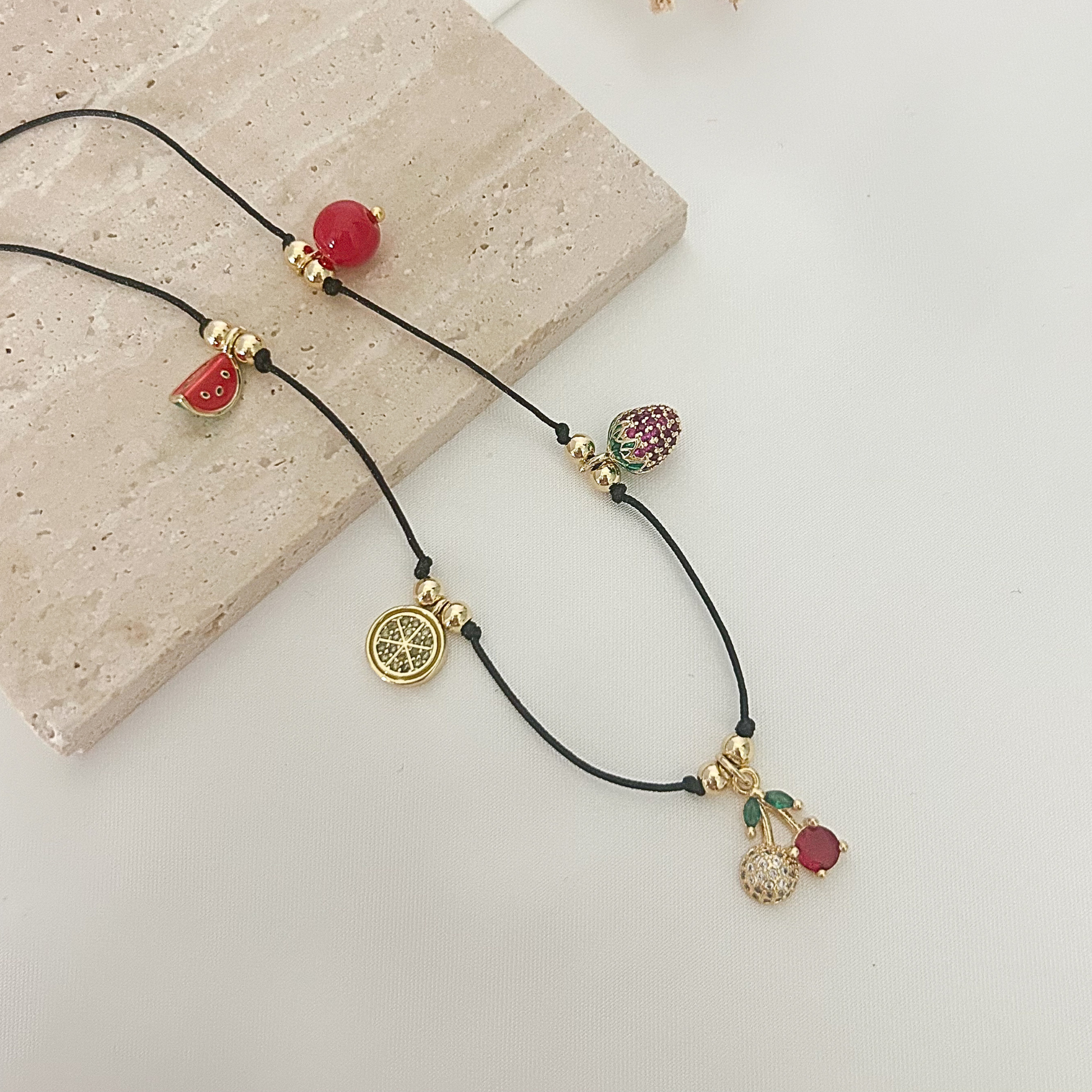 Playful charm necklace featuring a black cord, gold-tone details, and colorful fruit-inspired charms including cherries, strawberry, watermelon, and citrus slice.