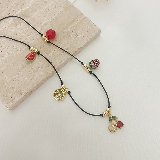 Playful charm necklace featuring a black cord, gold-tone details, and colorful fruit-inspired charms including cherries, strawberry, watermelon, and citrus slice.