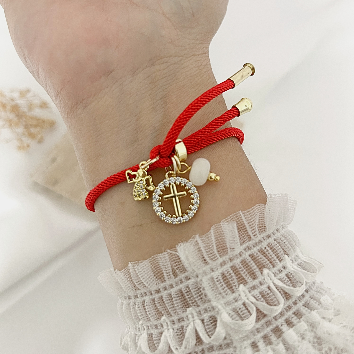 Red adjustable bracelet with zirconia cross, angel, and white bead charms worn on the wrist