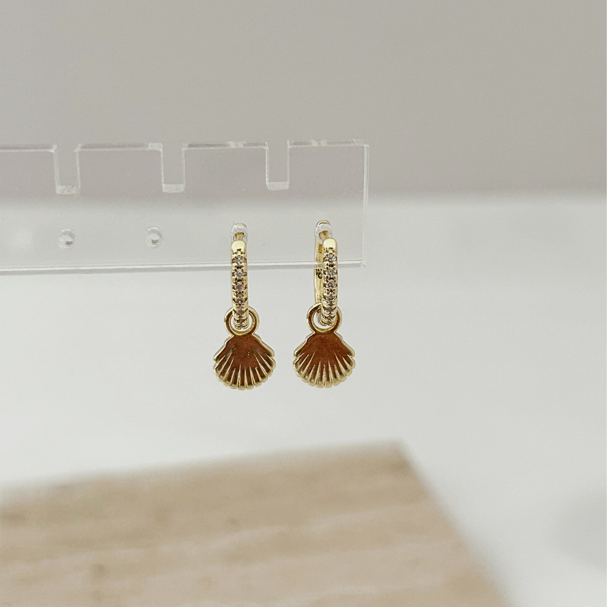 A pair of gold dangle earrings with a scallop-shaped charm and small clear stones, hanging from a clear display stand against a soft neutral background.