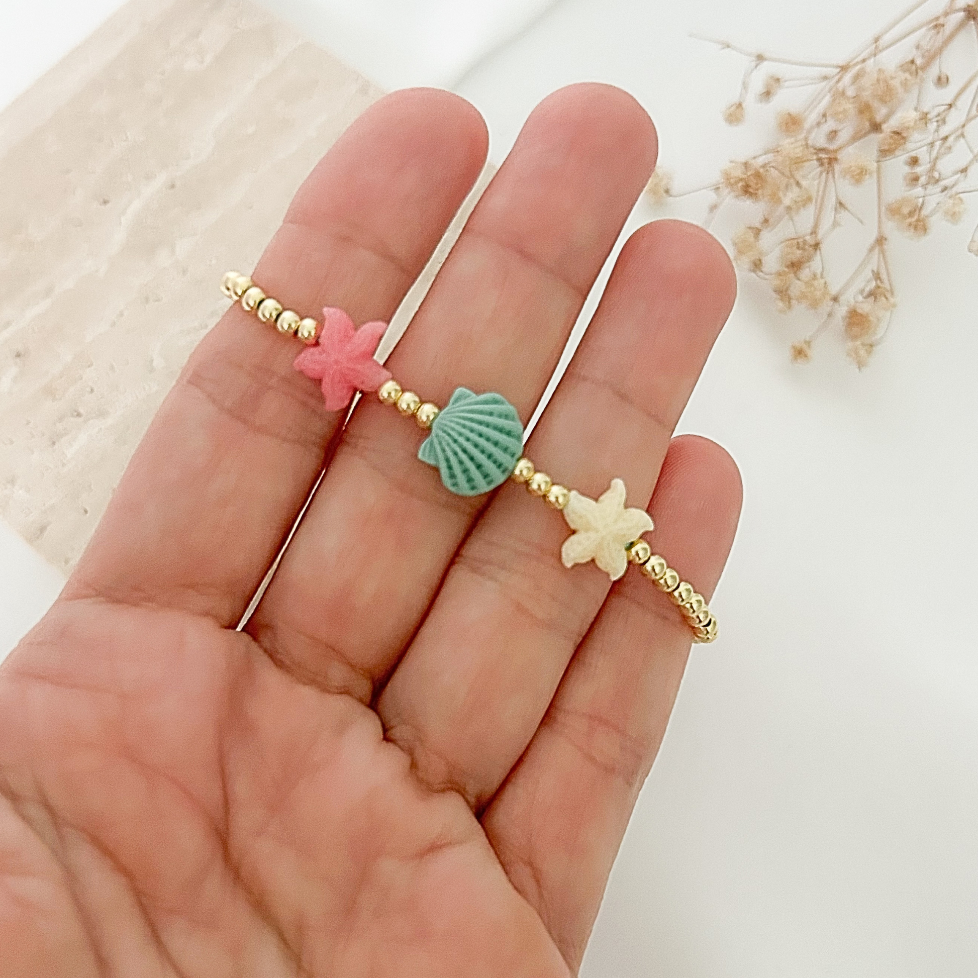 Close-up of an adjustable beach-themed bracelet featuring gold-tone beads with pastel starfish and seashell accents in pink, mint green, and ivory, displayed in hand against a soft neutral background.