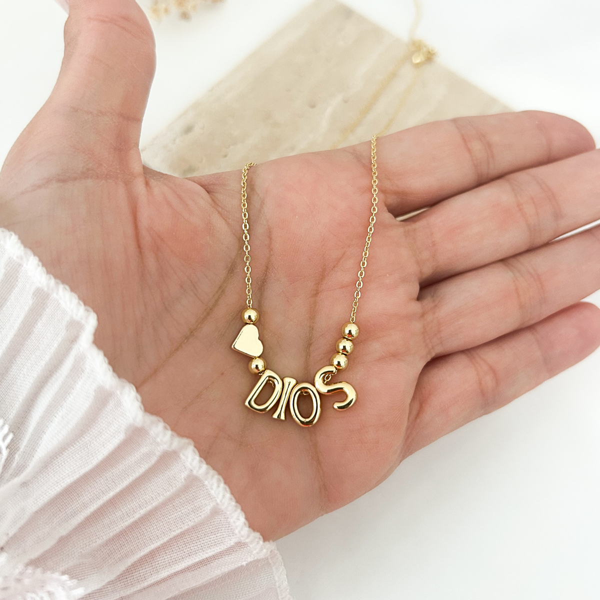 A gold necklace with a 'DIOS' letter pendant, a small heart charm, and spacer beads, displayed on an open hand.