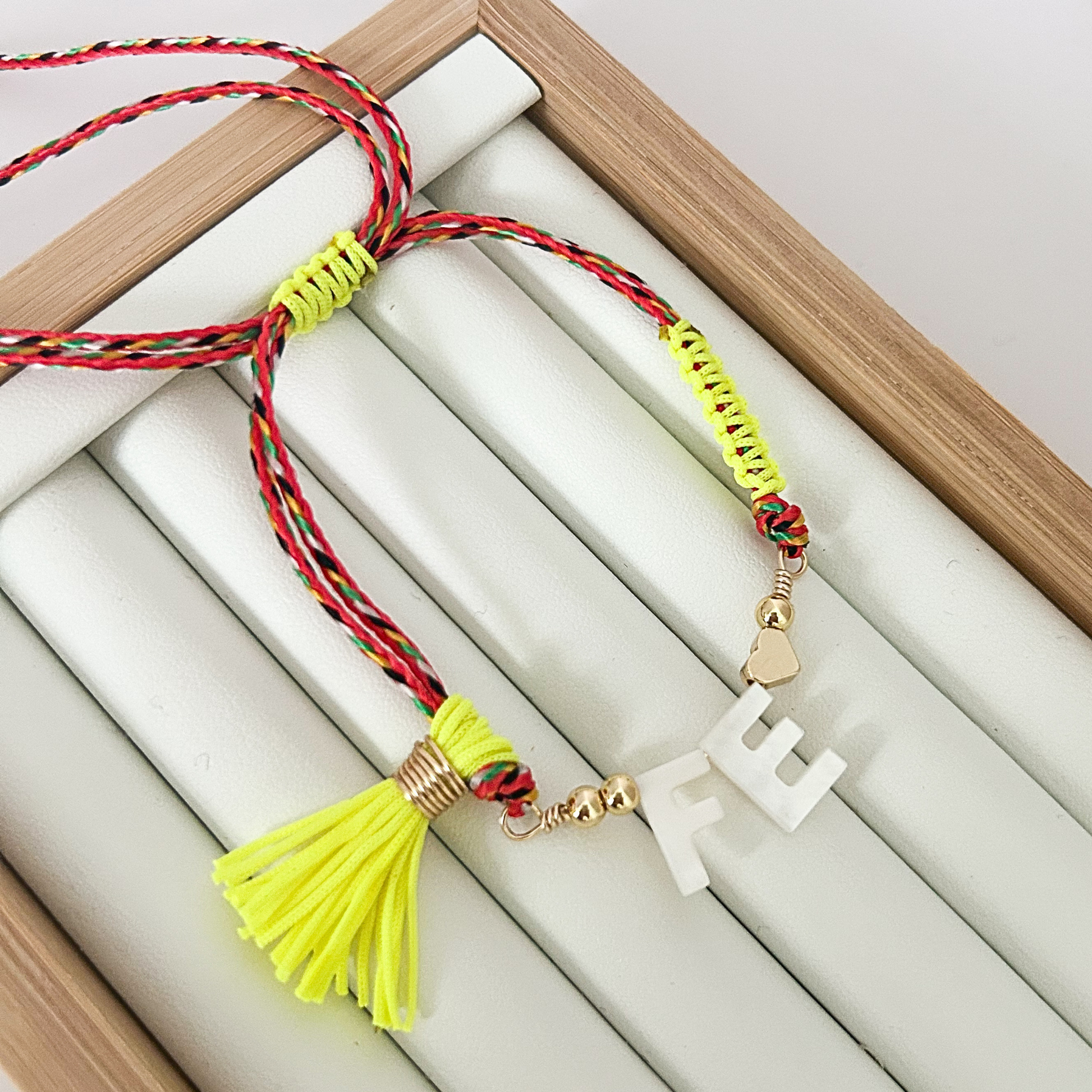 Adjustable multicolor cord faith bracelet with neon yellow tassel and white Fe letter gold charm on jewelry display