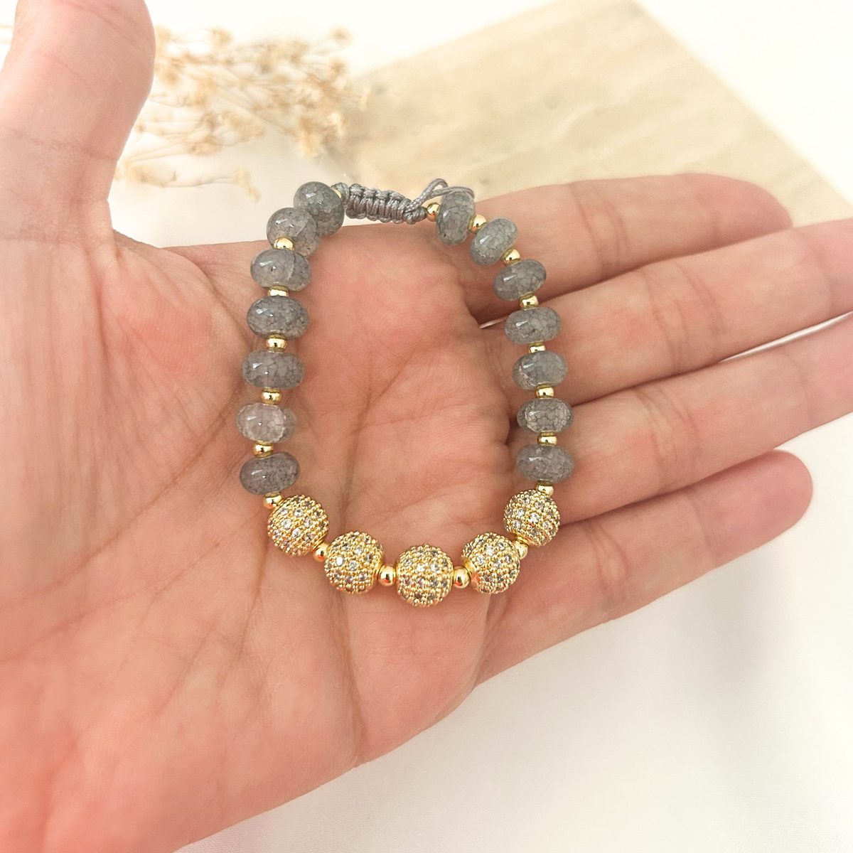 A beaded bracelet with gray stone beads, gold spacer beads, and three textured gold dome beads, displayed on a person's hand.