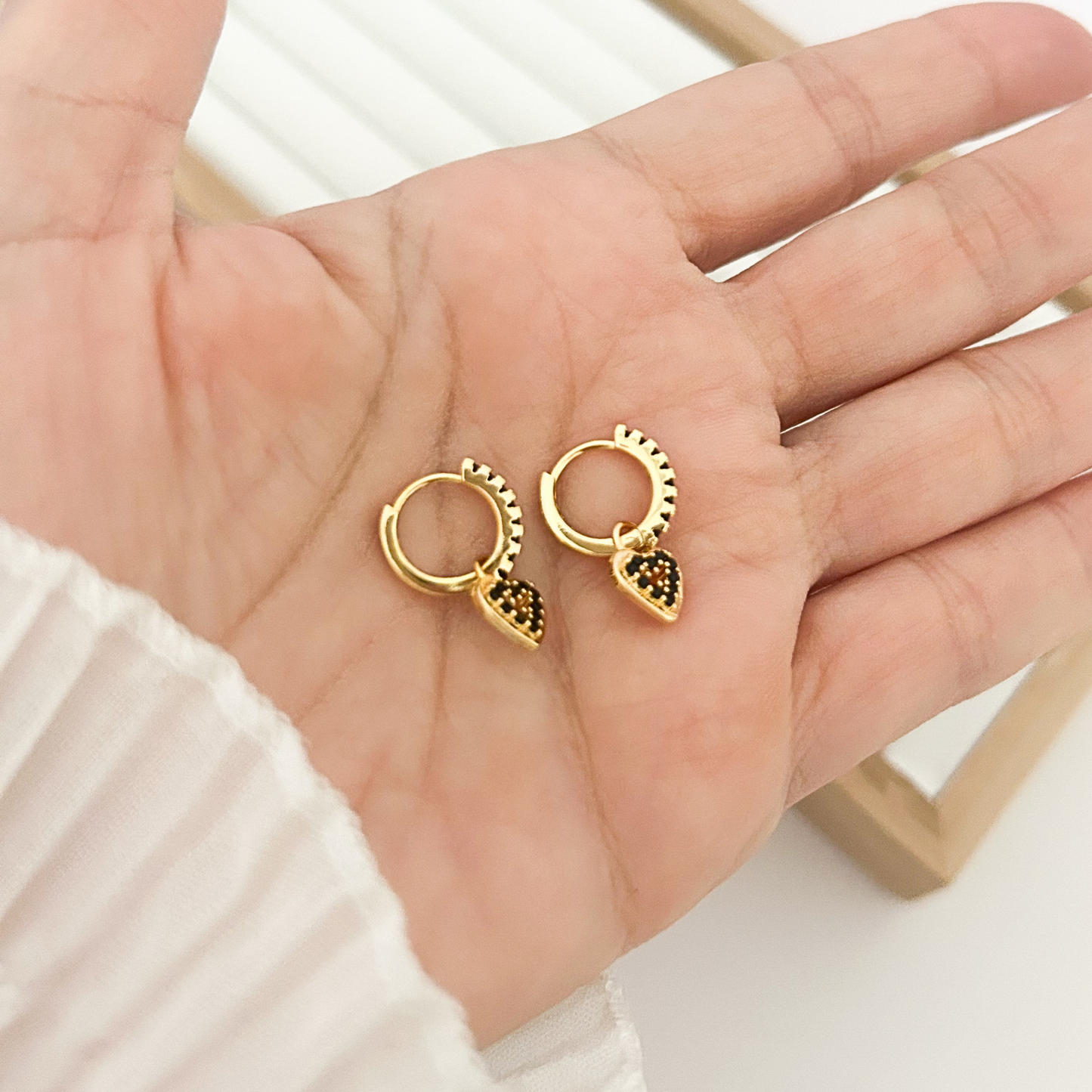 Pair of gold huggie hoop earrings with black pavé heart charm held in palm
