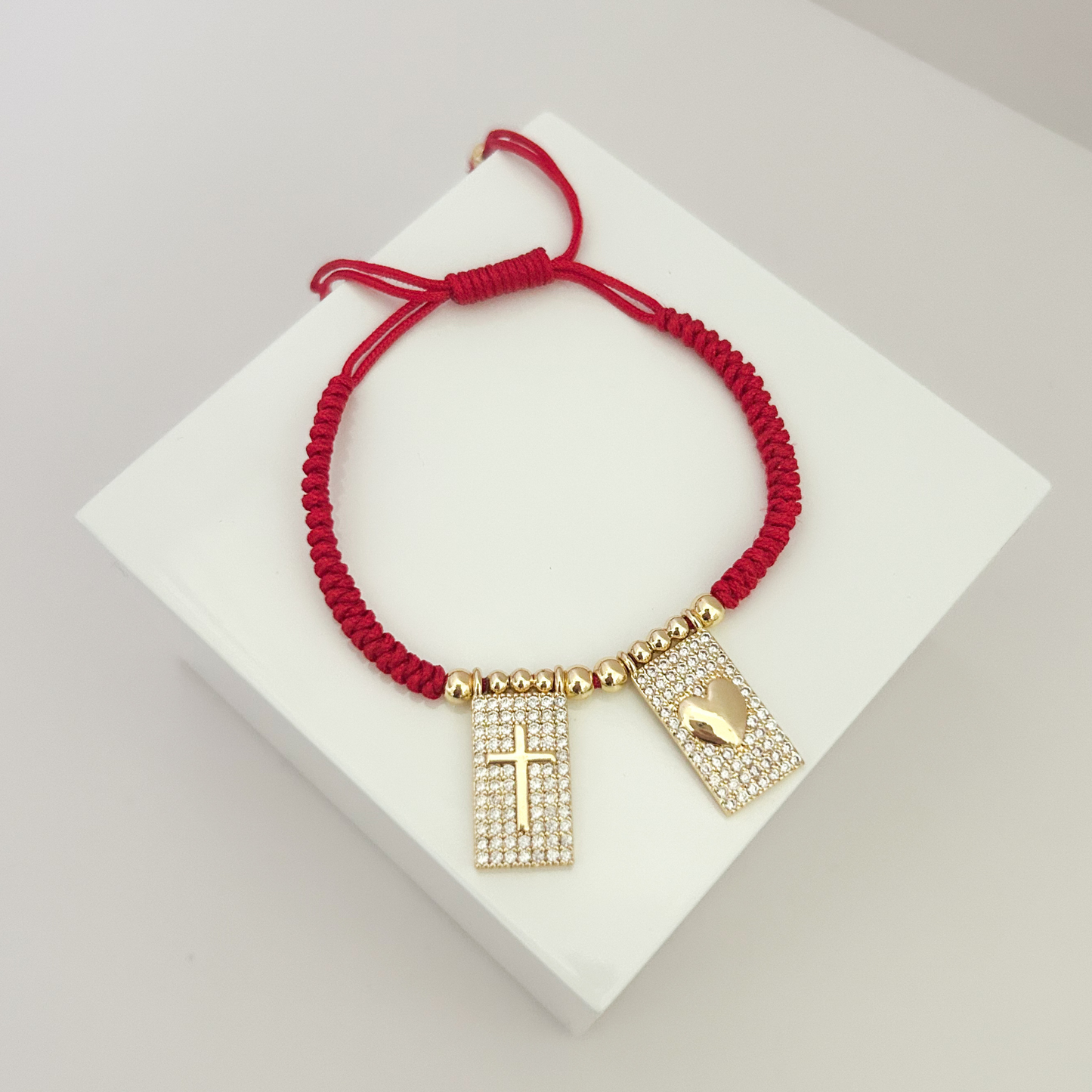 Adjustable red cord bracelet featuring gold-tone accents and sparkling rectangular charms with a cross and heart design, displayed on a minimalist white stand.