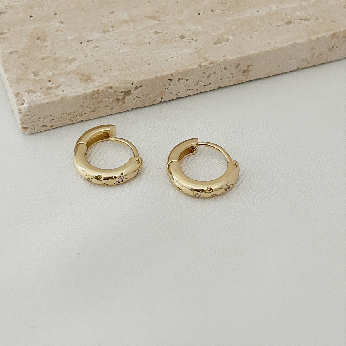 A pair of small gold huggie hoop earrings with a textured, hammered finish, displayed on a light gray surface with a beige stone tile in the background.