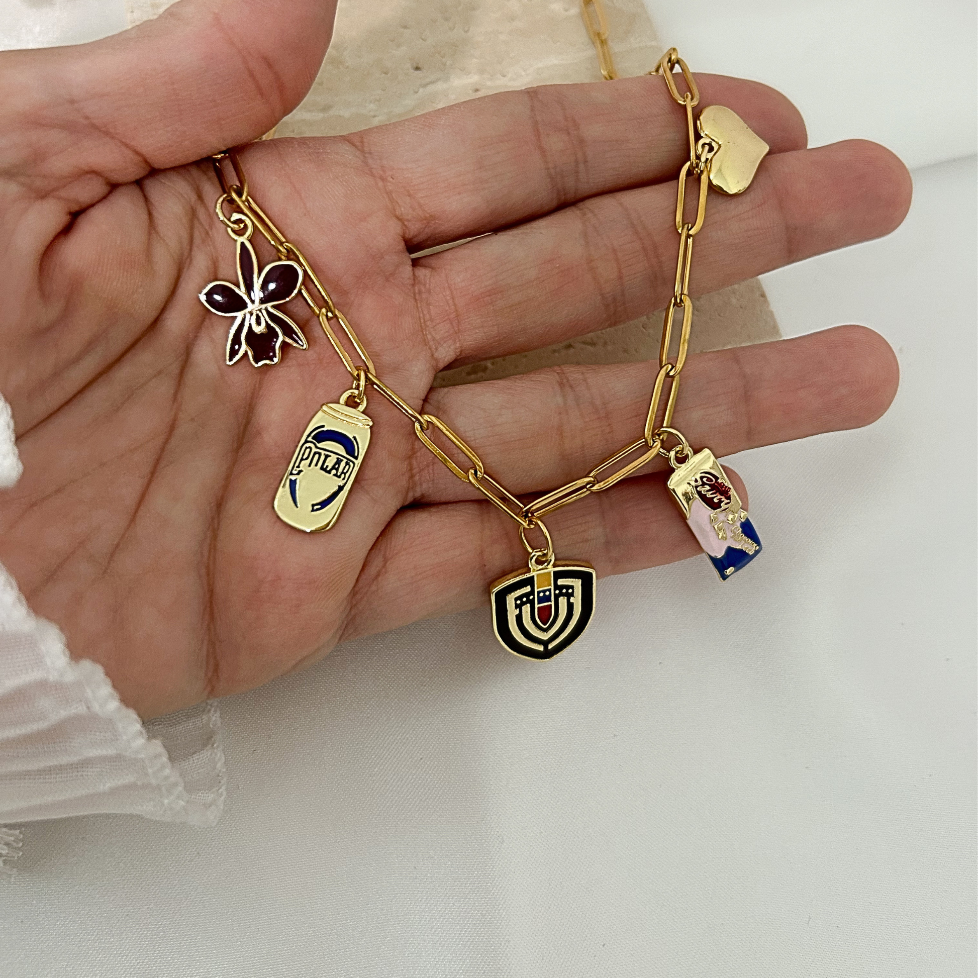 Gold paperclip charm necklace with iconic Venezuelan-inspired pendants, including an orchid, Polar beer can, Savoy chocolate bar, Venezuelan shield, and heart charm, shown in hand to highlight its size and unique details.