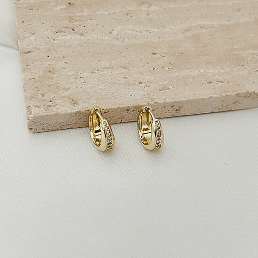A pair of small gold huggie hoop earrings with clear stones, displayed on a light beige stone slab over a white surface.