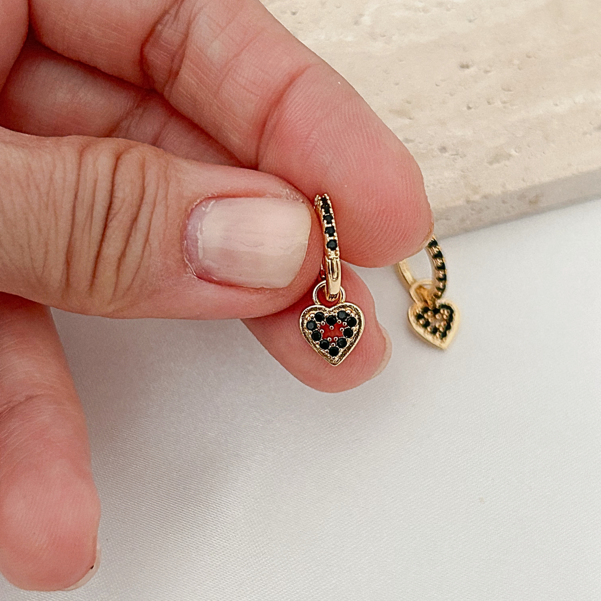 A close-up of a hand holding a small gold heart charm with black and red stones, with another matching charm visible in the background.