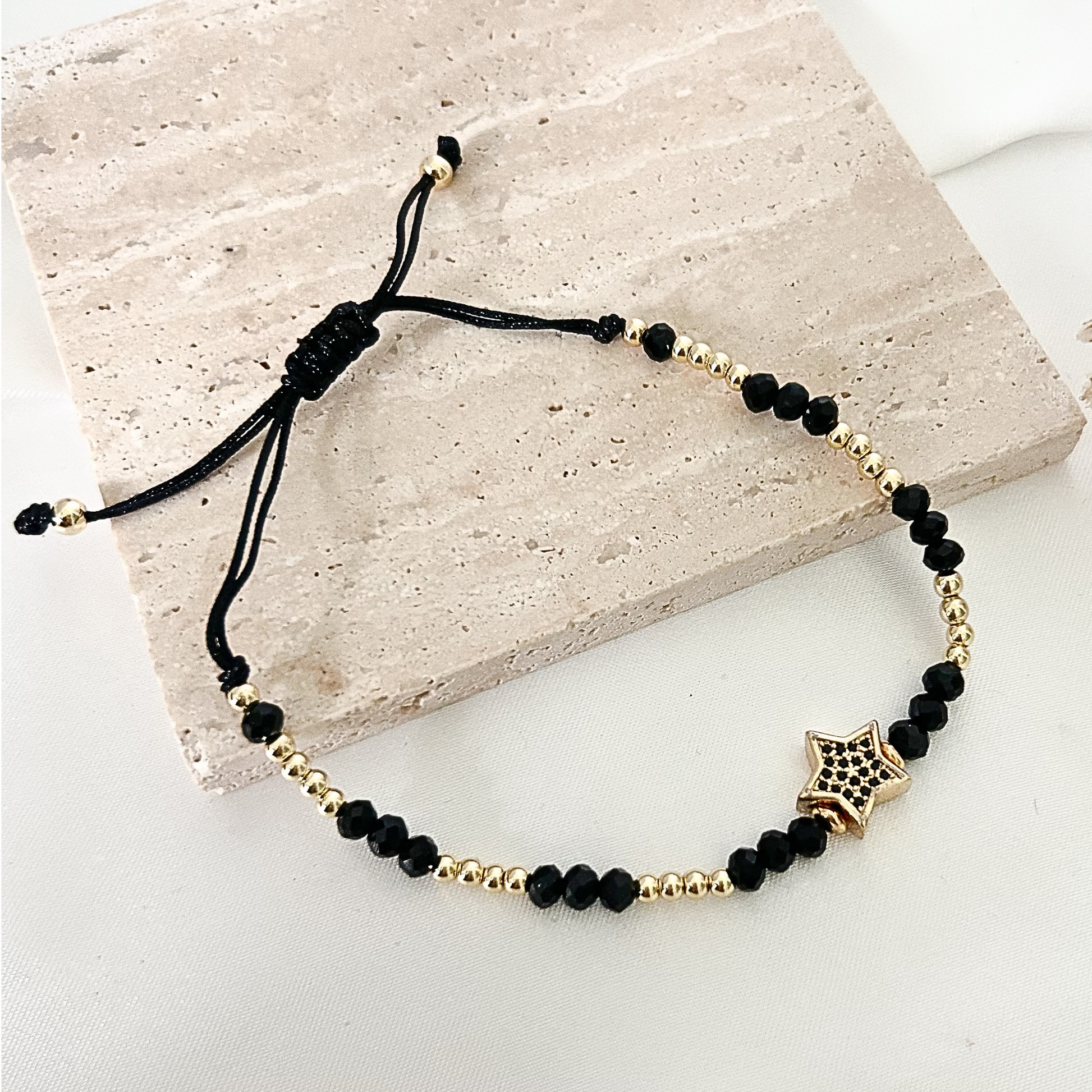 Adjustable bracelet crafted with black faceted beads and polished gold-tone spacer beads, centered with a decorative star charm, displayed on a neutral stone surface.
