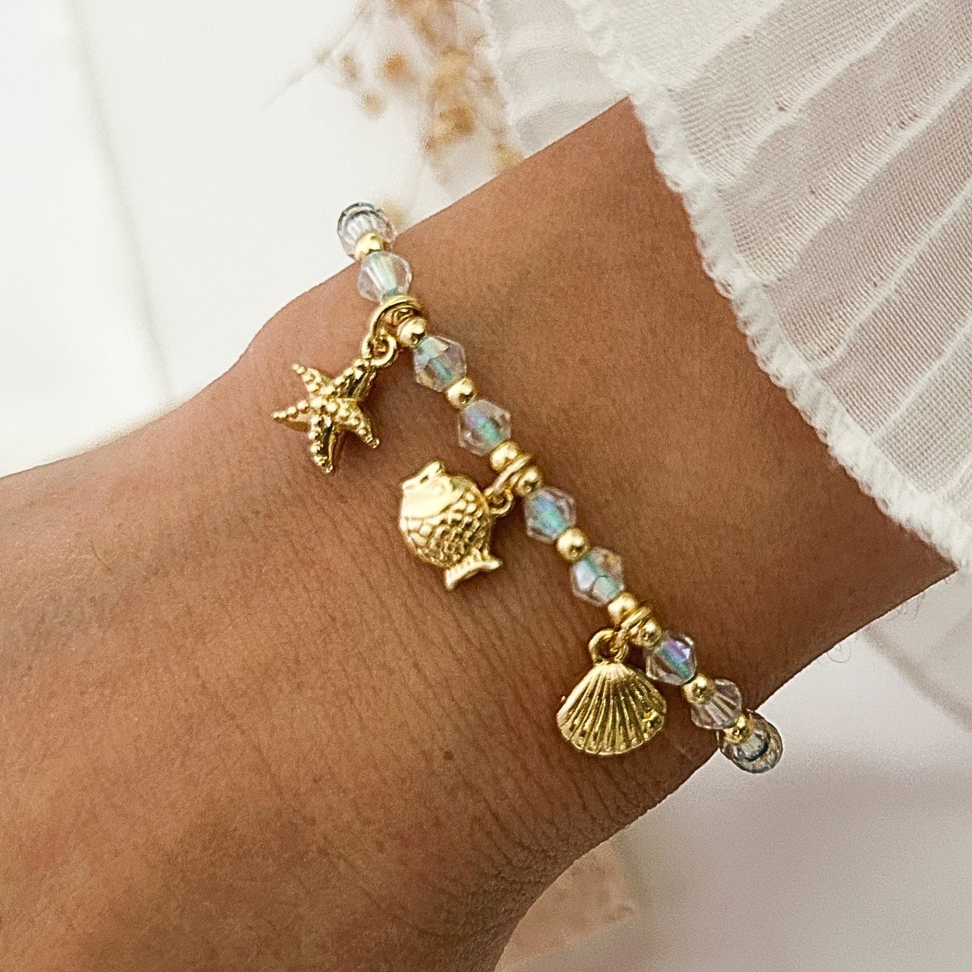 Beach-inspired bracelet with iridescent crystal beads, gold-tone accents, and dangling starfish, fish, and seashell charms, worn on the wrist with a white ruffled sleeve.