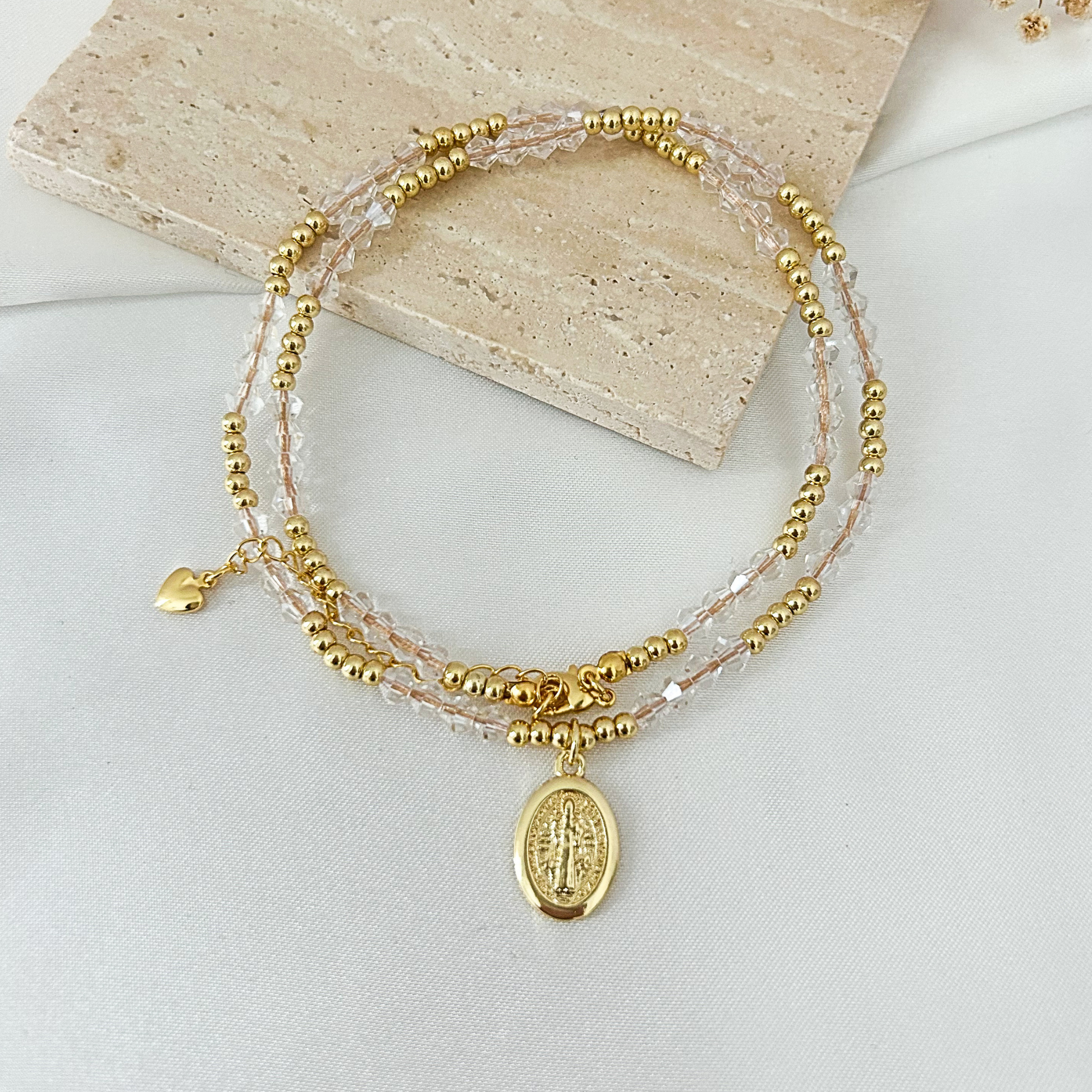 A delicate multi-strand beaded bracelet with gold-tone beads, clear faceted beads, a small gold heart charm, and an oval gold-tone religious medallion pendant, displayed on a light fabric surface with a stone tile prop.