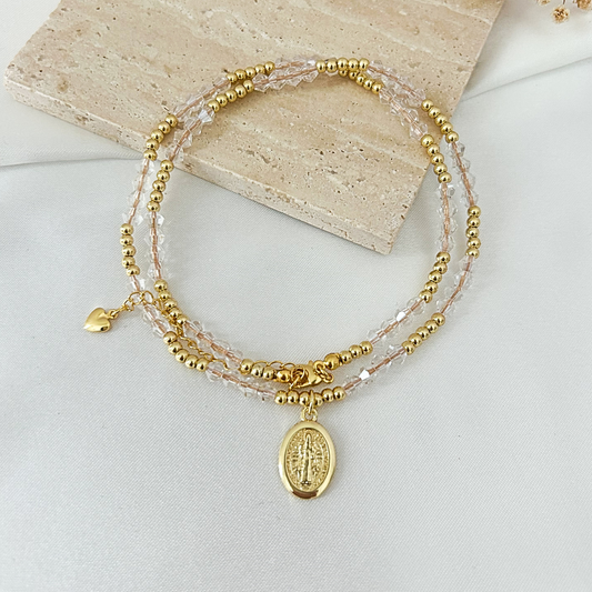 A delicate multi-strand beaded bracelet with gold-tone beads, clear faceted beads, a small gold heart charm, and an oval gold-tone religious medallion pendant, displayed on a light fabric surface with a stone tile prop.